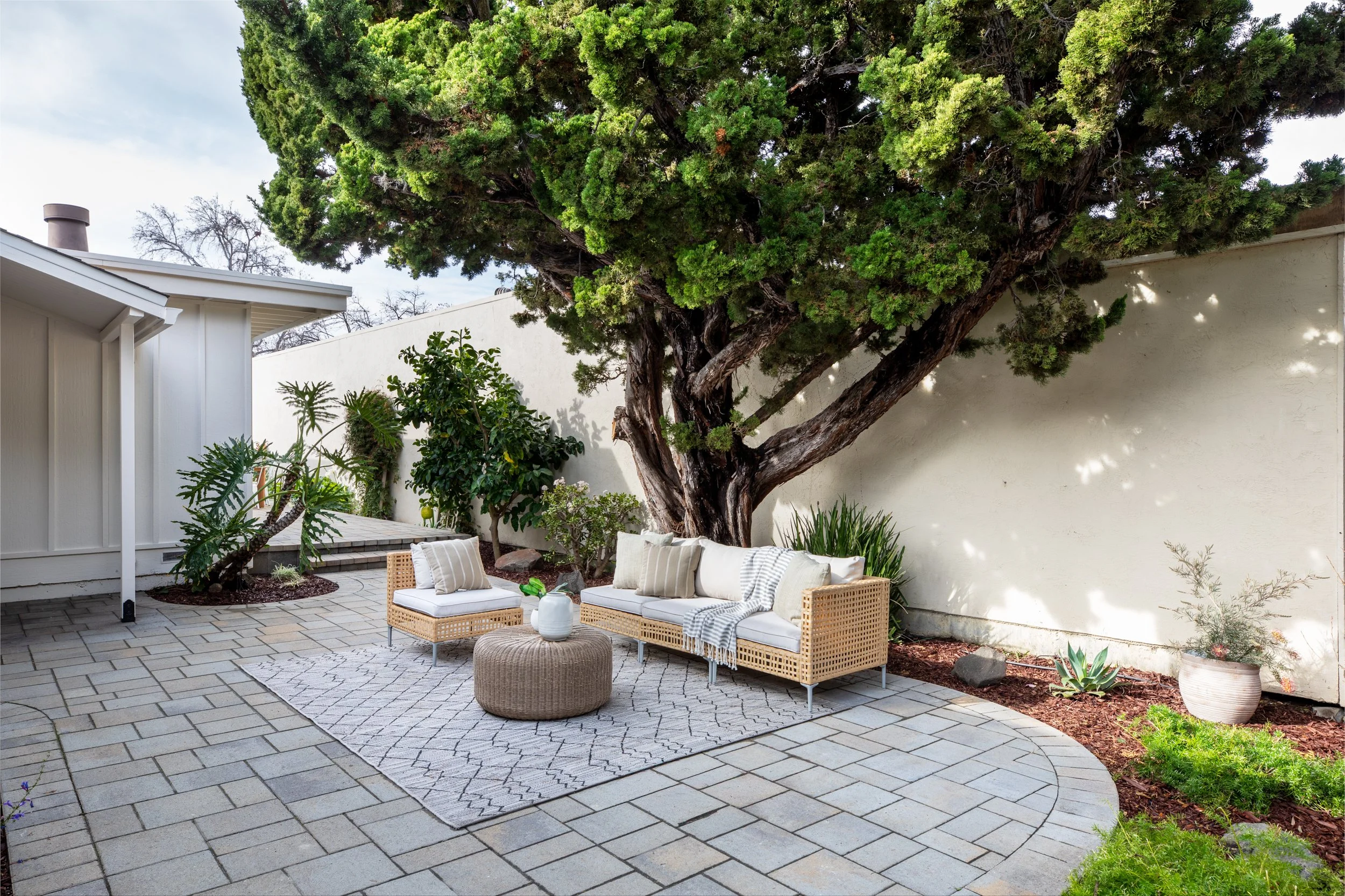 Outdoor patio with wicker sofa and matching chairs with cushions, a round ottoman, and a geometric-patterned rug, surrounded by potted plants and a large tree providing shade, with a white wall in the background.