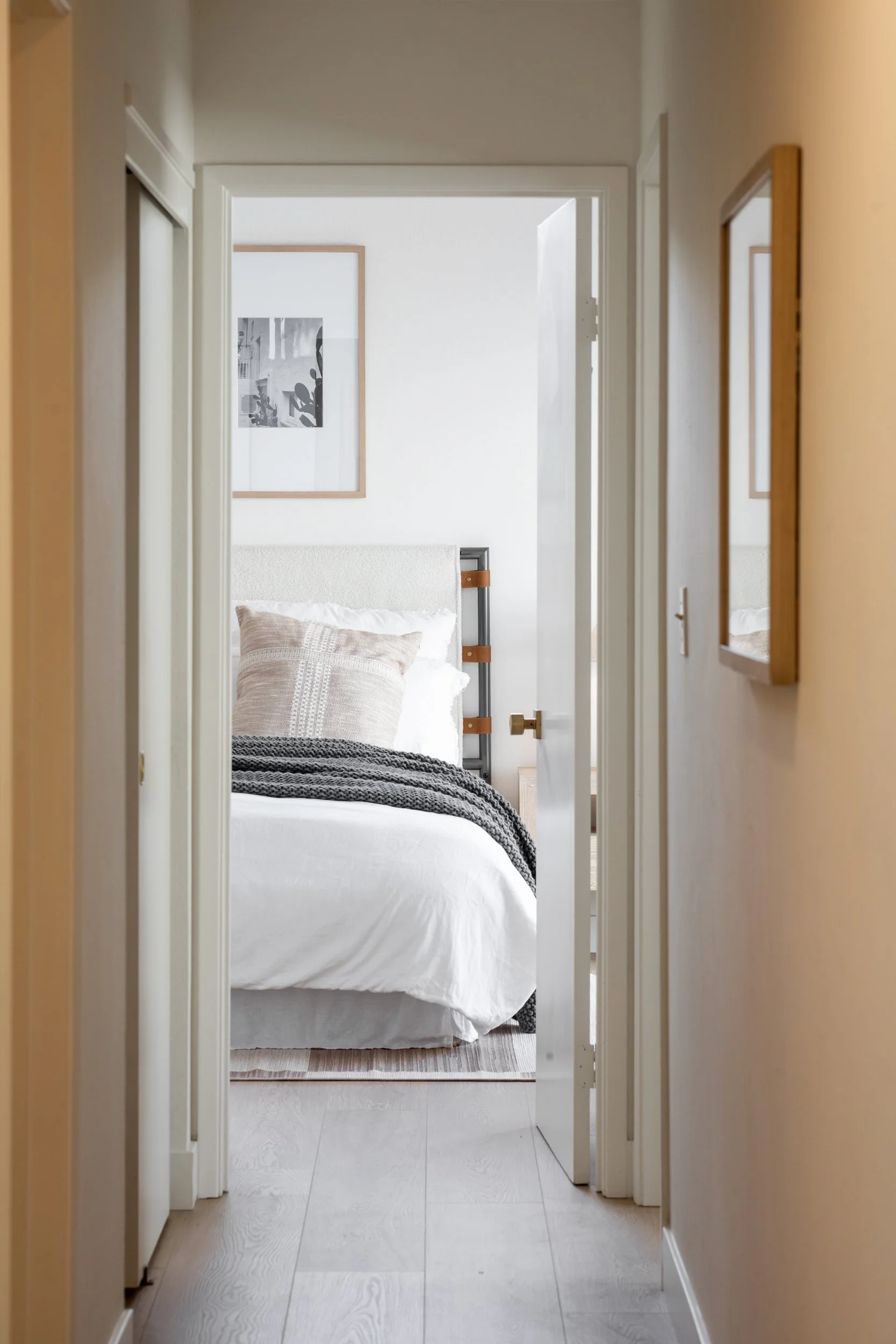 View of a neatly made bed with beige and black pillows and a gray knitted throw blanket, visible through a doorway in a minimalistic, modern bedroom.