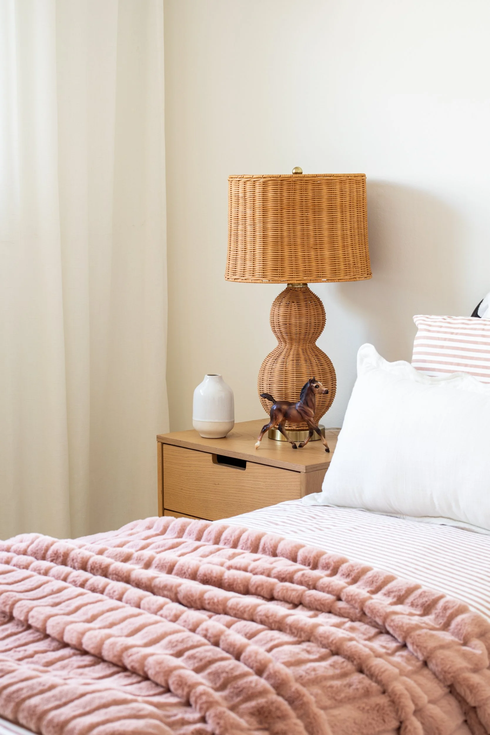 A bedroom with a wooden nightstand holding a rattan table lamp, a white vase, and a small horse figurine. The bed has white pillows, a striped pillow, and a pink fluffy blanket.
