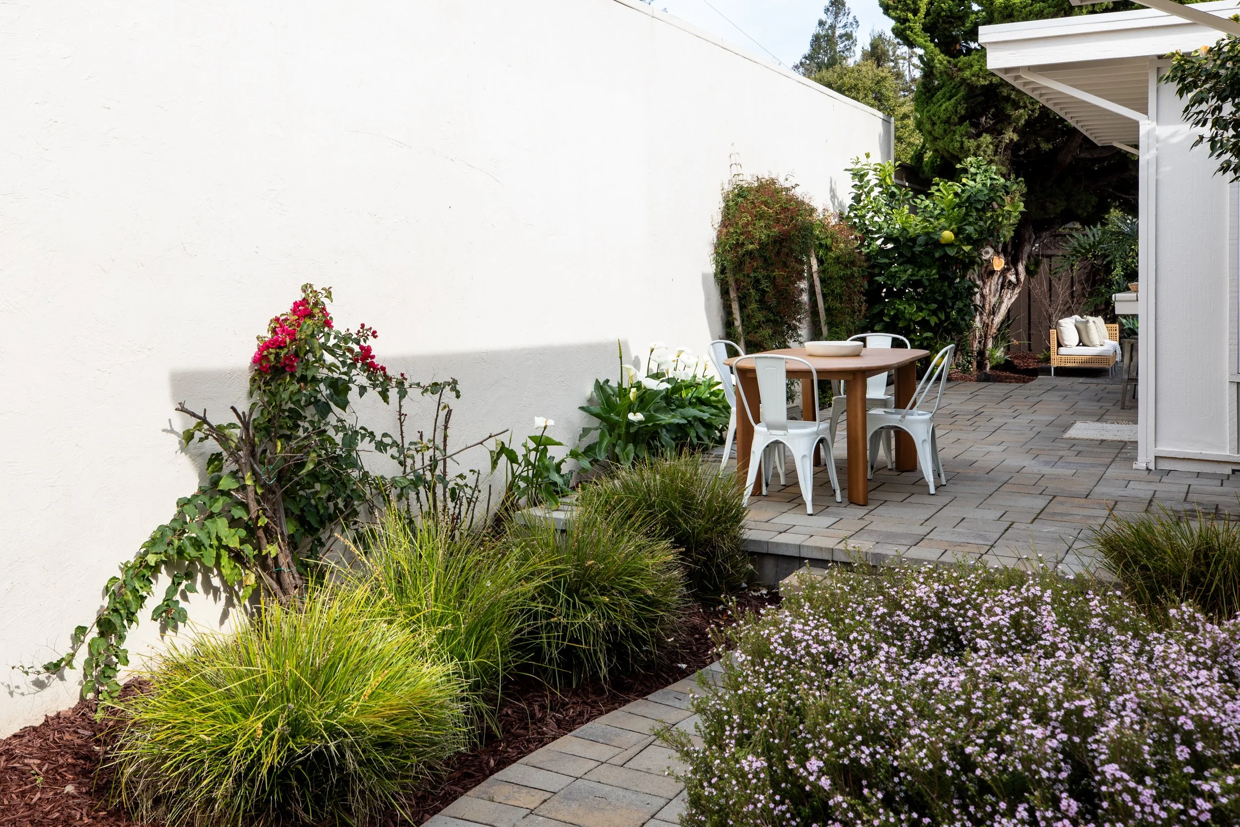 An outdoor garden patio with a white wall, flowering bushes, green plants, a wooden table with white metal chairs, a wicker bench with cushions, and trees in the background.