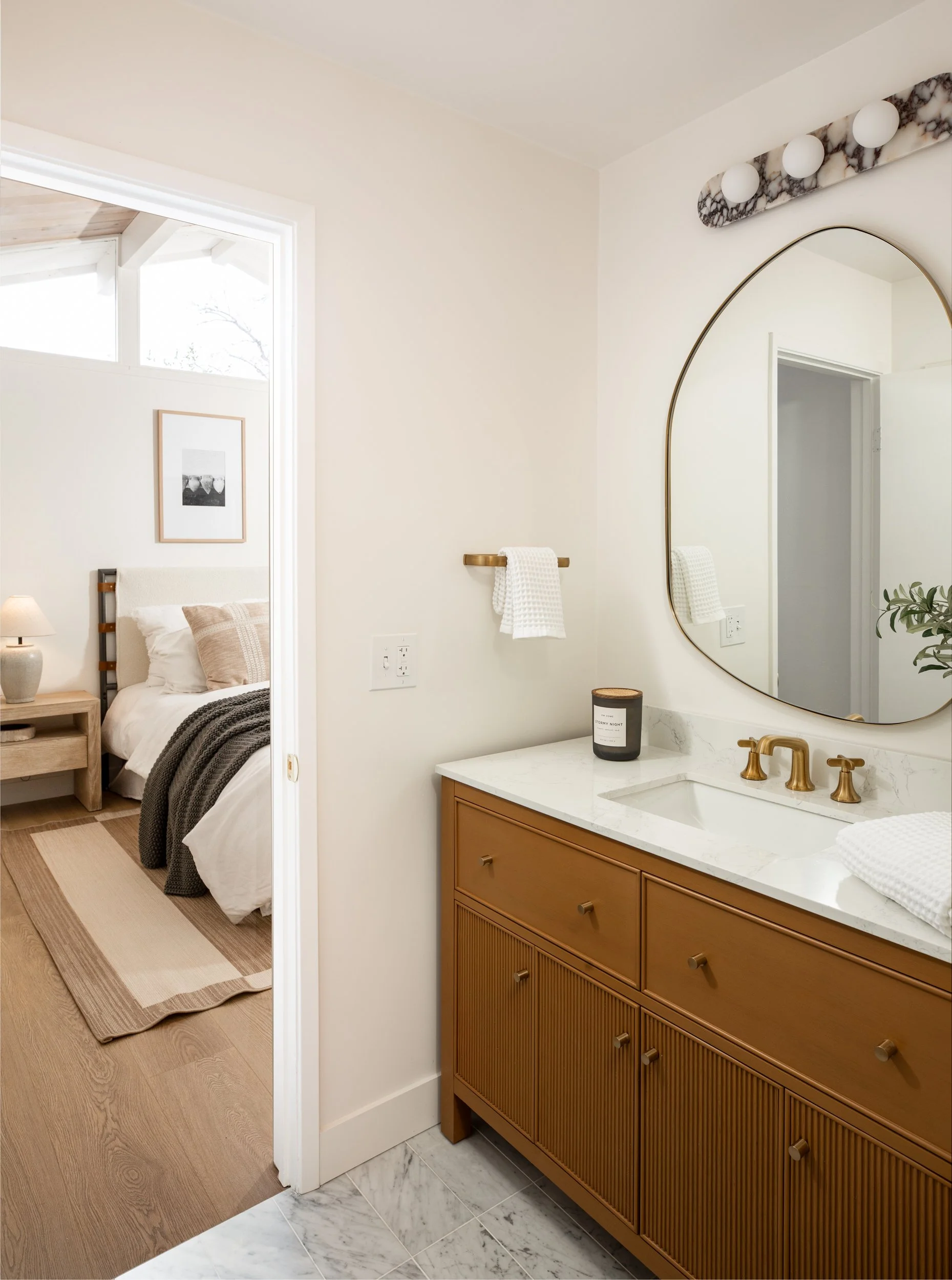 Bathroom with gold fixtures, a large mirror, a white marble countertop, and a wooden vanity. Visible part of a bedroom with similar decor.