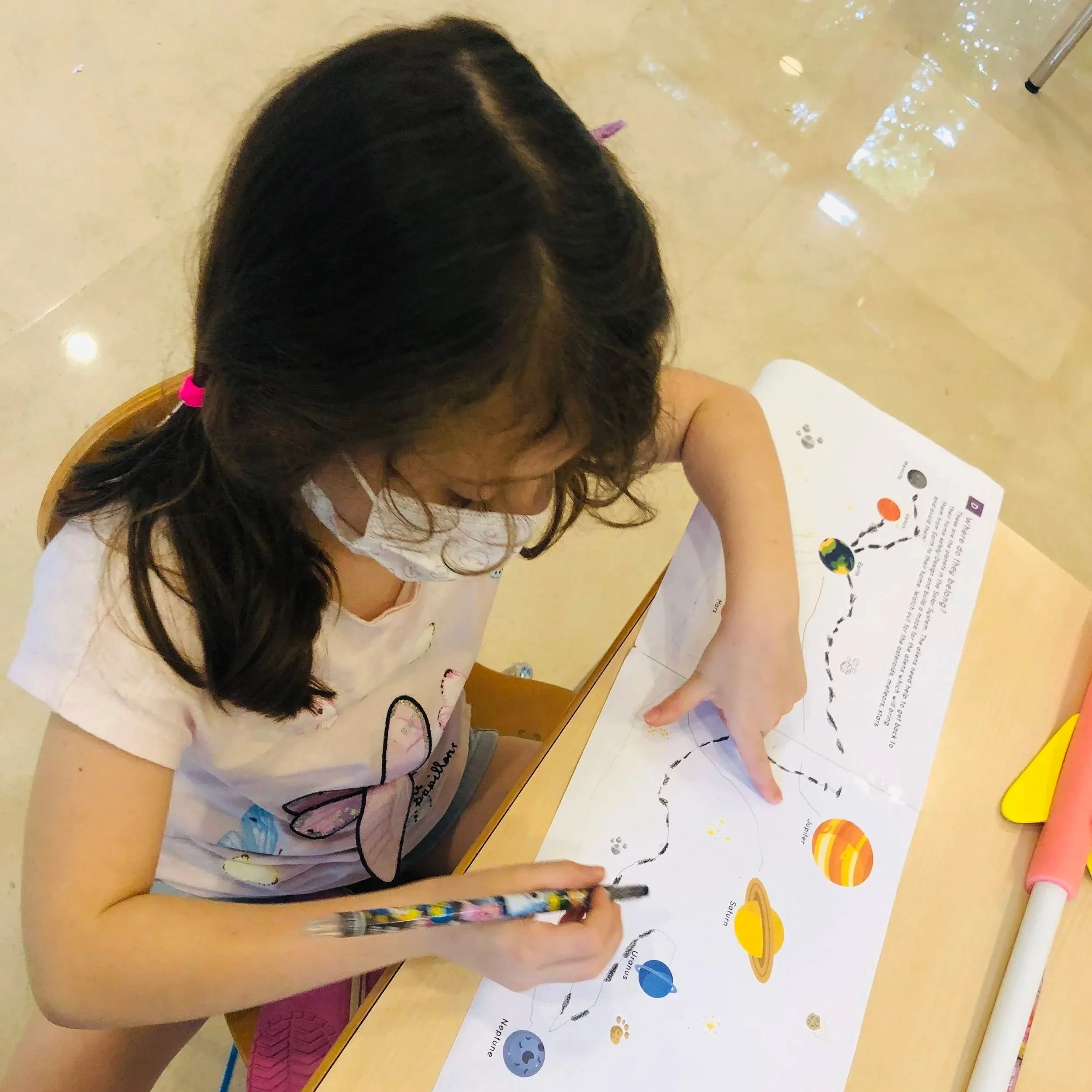 A young girl with long dark hair and a pink hair clip, wearing a white t-shirt with a graphic, is seated at a table drawing or writing on a sheet of paper with colorful planets and labels related to space.