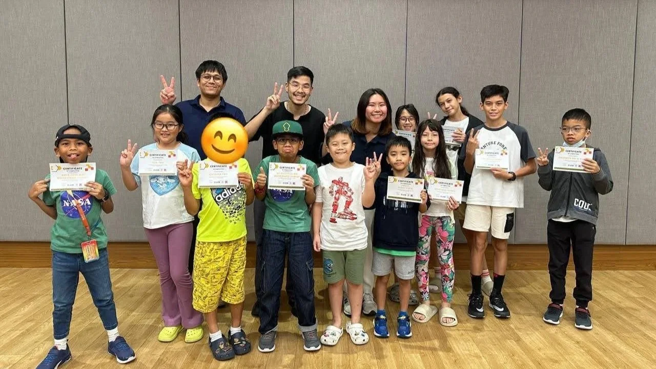 Group of children and adults posing indoors, some children holding certificates, others making peace signs, in front of a gray wall.