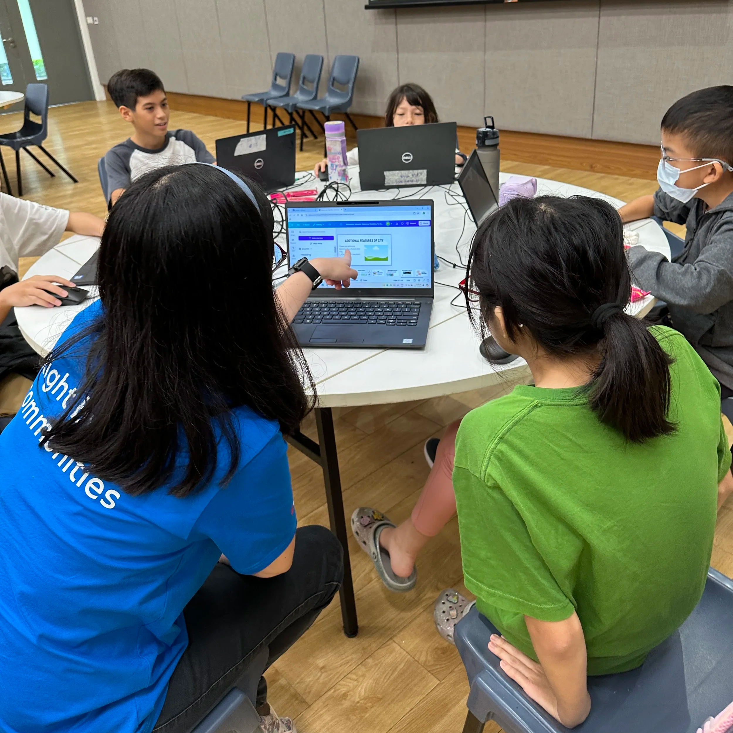 Children sitting at a round table with laptops, engaging in a group activity in a classroom setting.