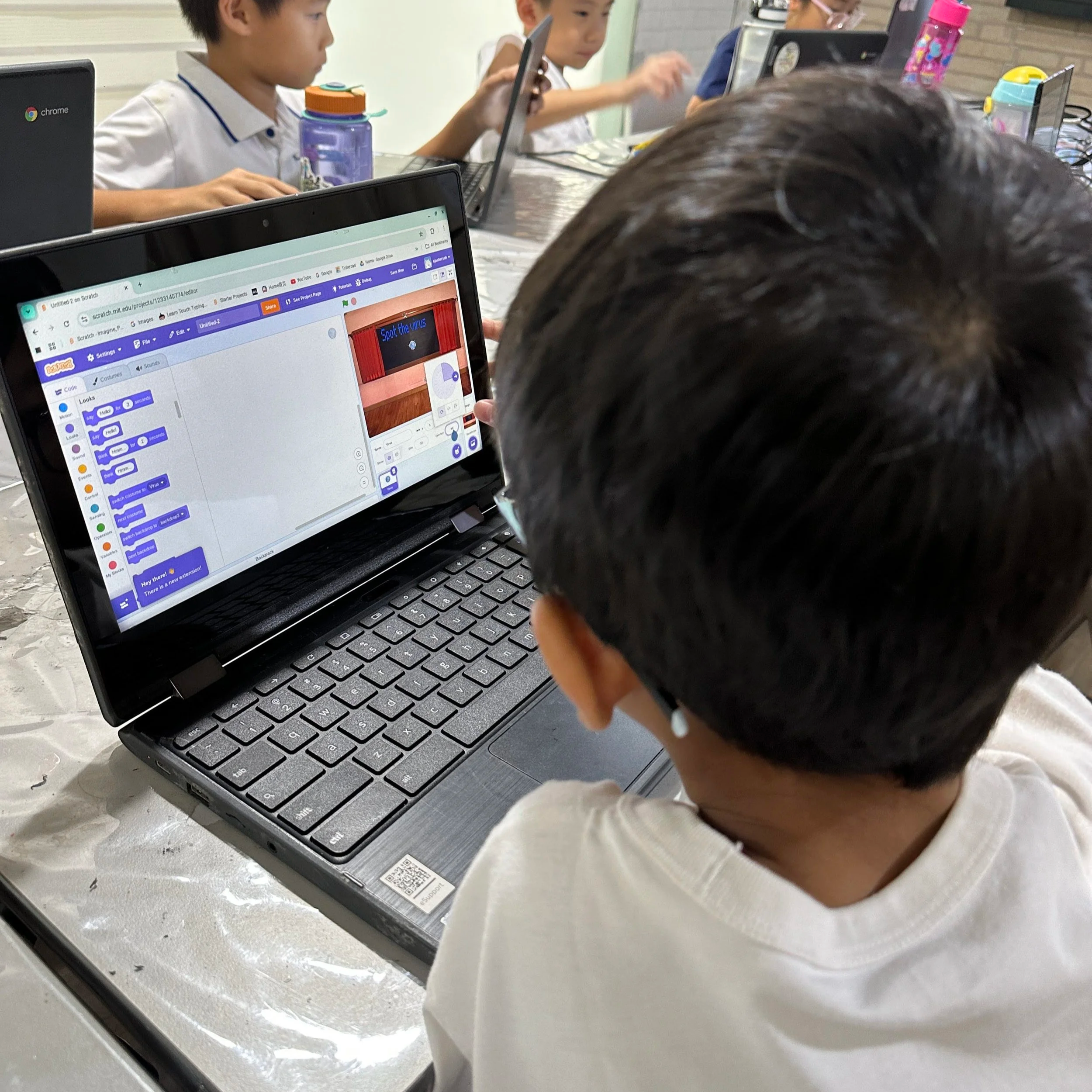 A group of children in a classroom, focused on their laptops, with one child in the foreground working on a computer displaying a coding project, 'Spot the virus'.