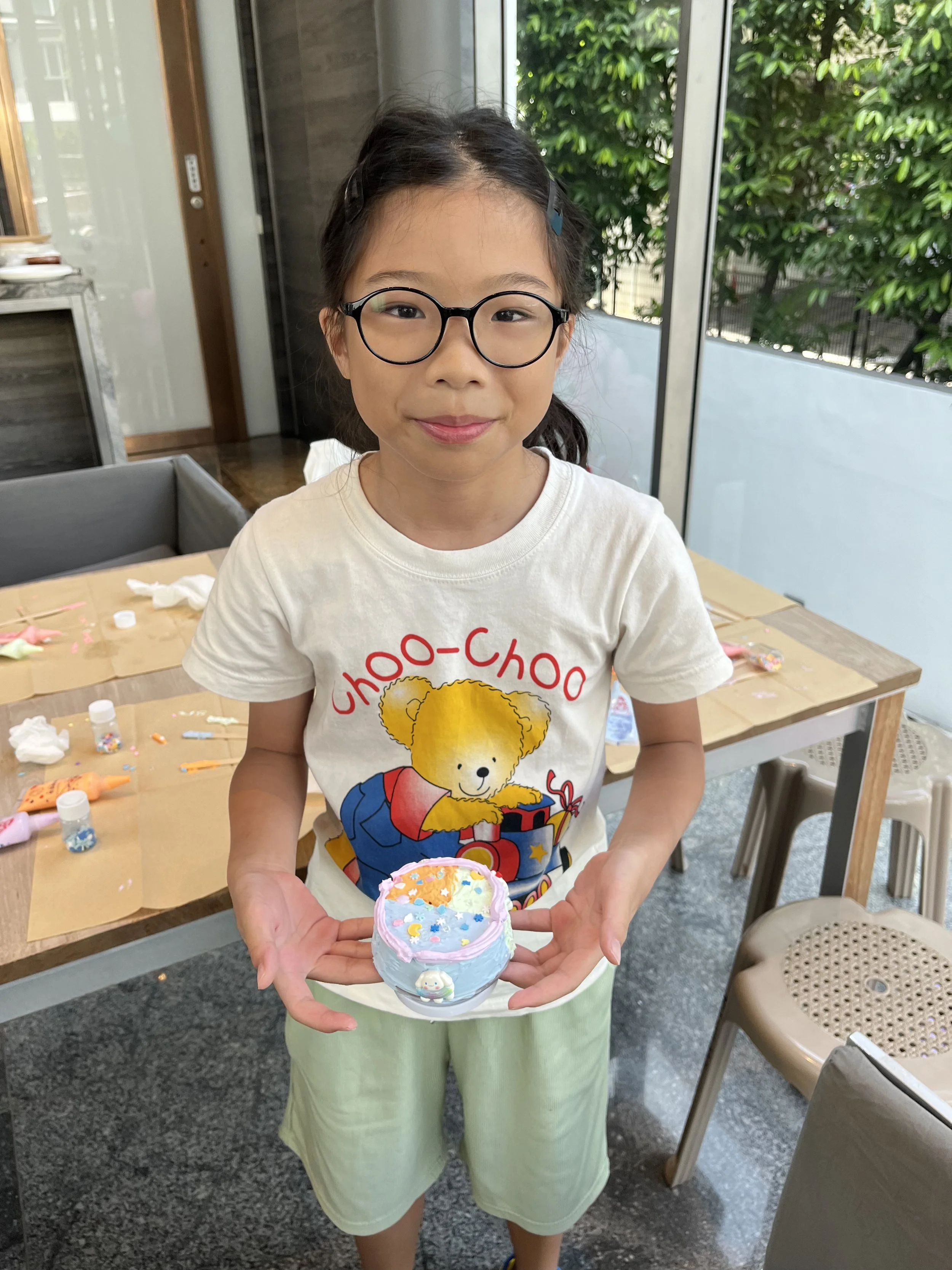 A young girl with glasses holding a decorated container, standing in a room with tables and chairs, with arts and crafts items on the table behind her.