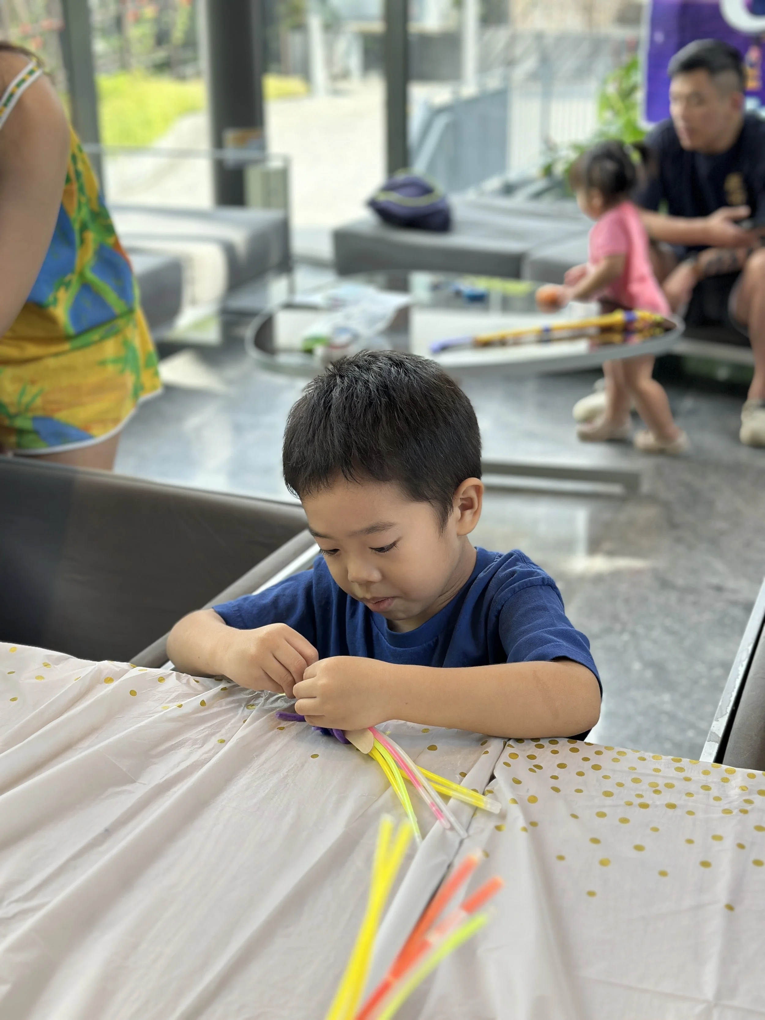 A young boy is sitting at a table, making crafts with colorful glow sticks, with other children and adults in the background in a bright, modern indoor space.