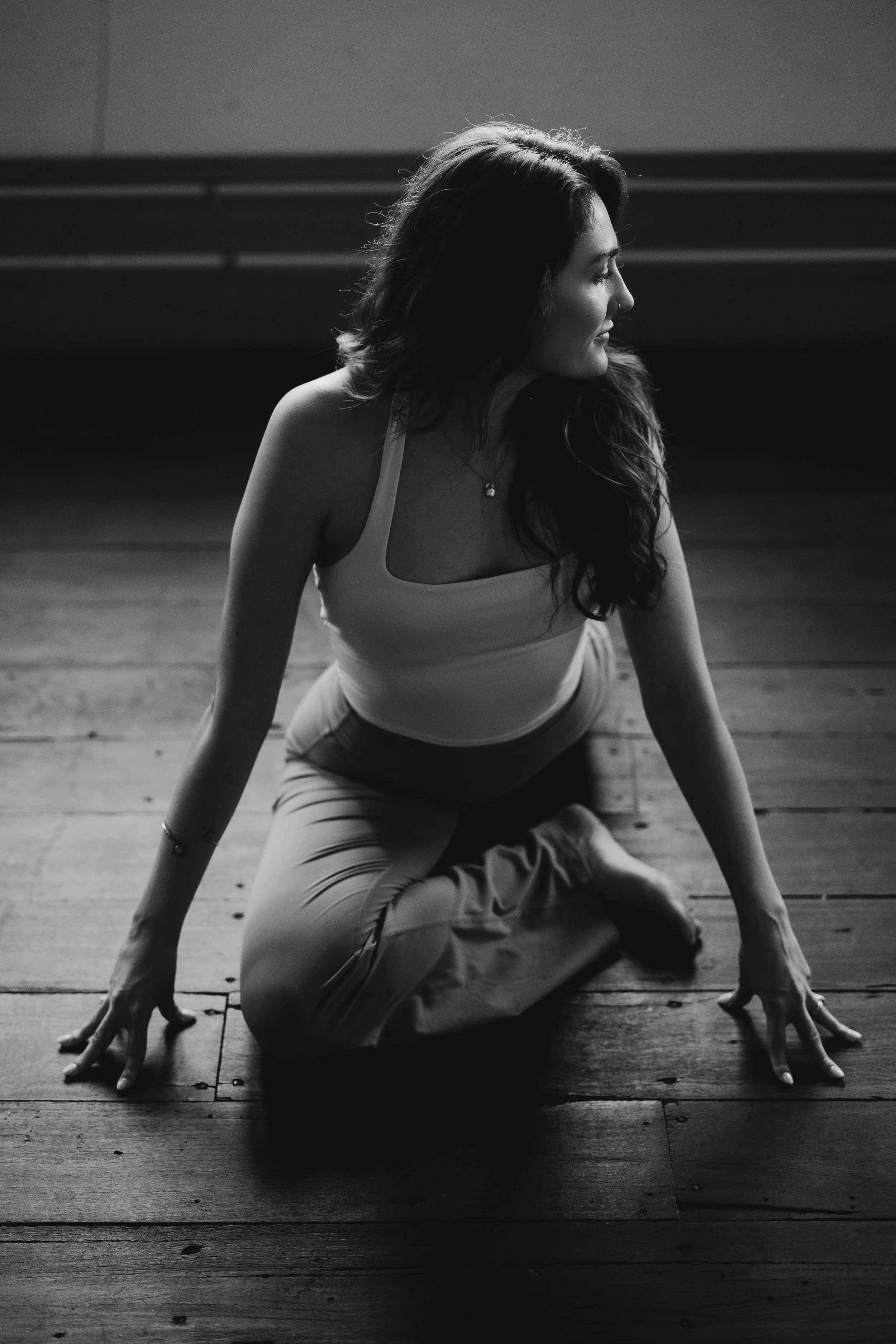A woman in yoga pants and a tank top is practicing yoga on a wooden floor, in a room with minimal lighting, captured in black and white.