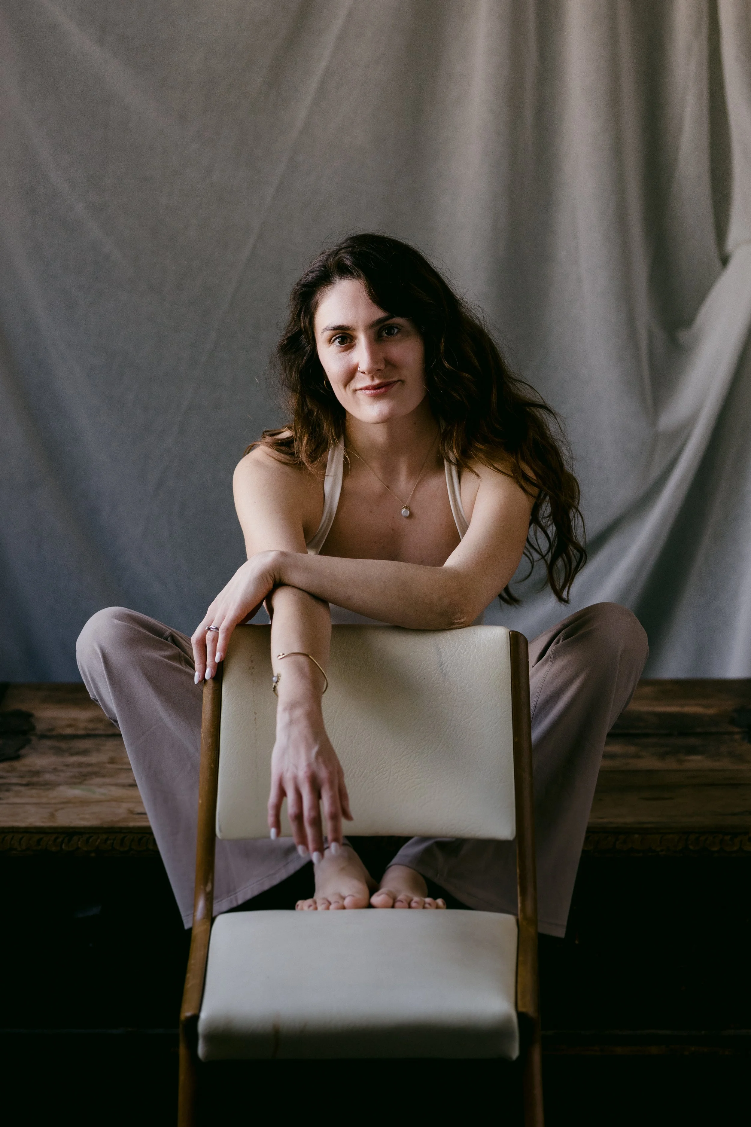 A woman with long dark hair sitting behind a white chair, with her arm resting on the backrest, and her foot on another chair in front of her. She is smiling softly and wearing a pearl necklace and beige pants. The background is a draped gray fabric.