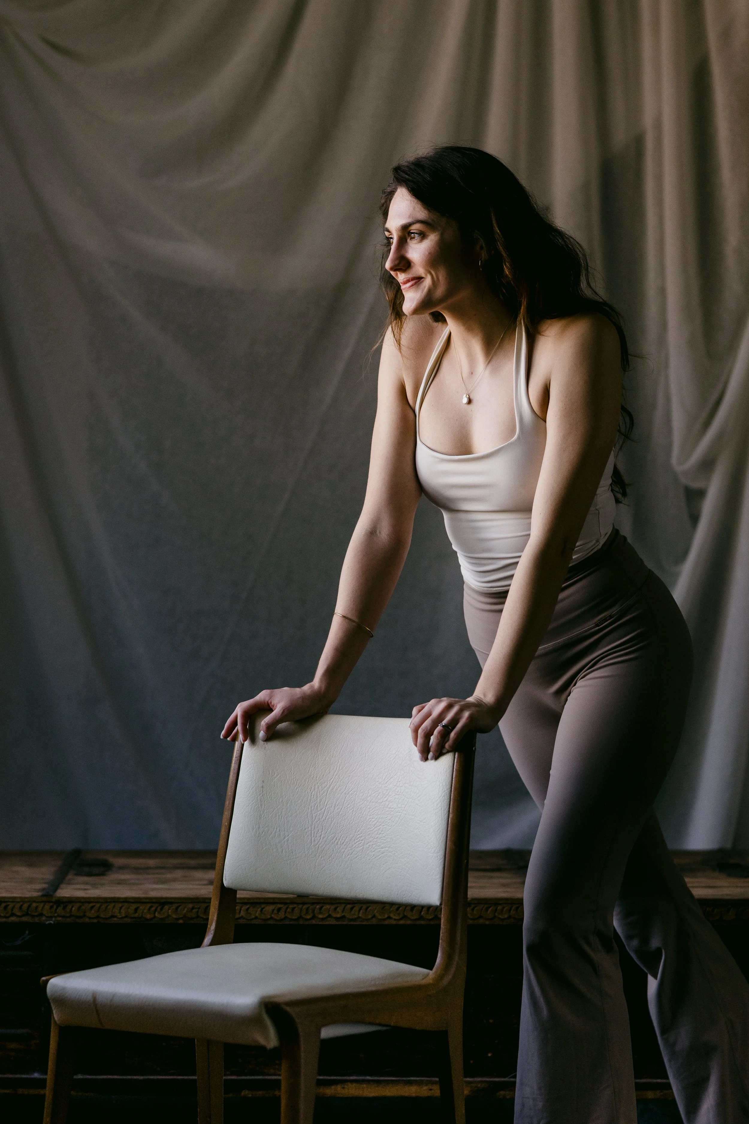 A woman with dark hair in a white tank top and gray pants, smiling, leaning on a chair with a plain, draped gray backdrop behind her.