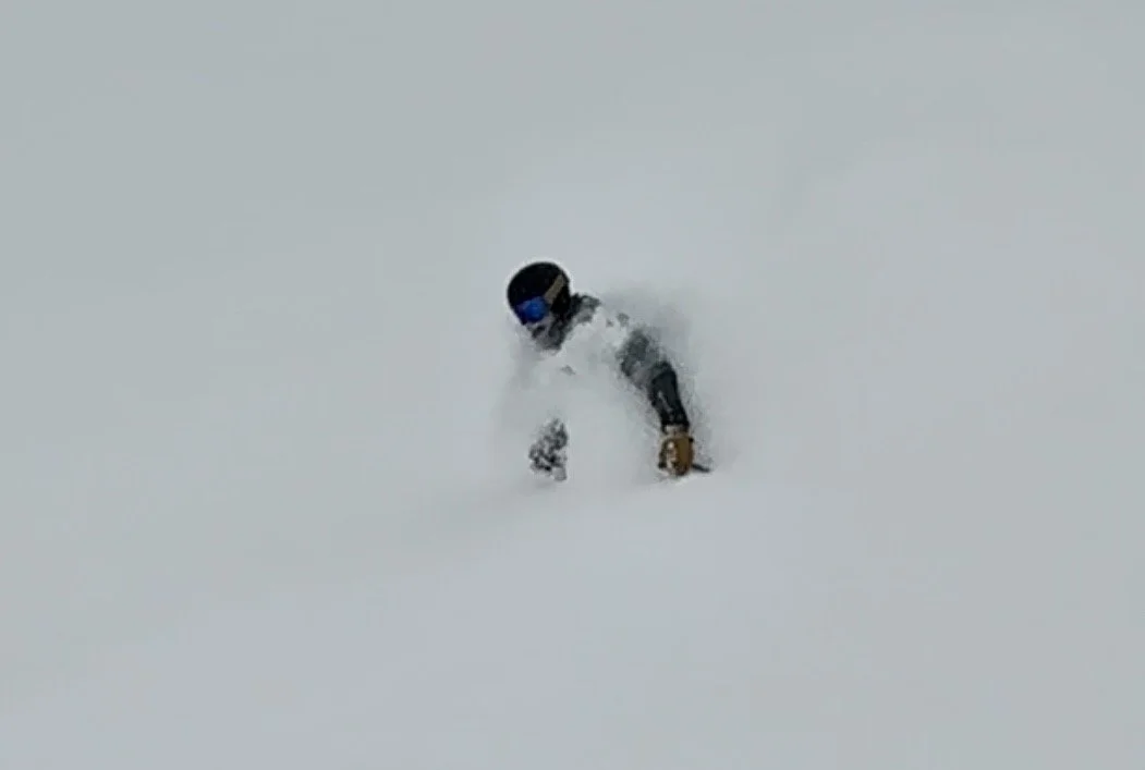 A person wearing a black helmet, goggles, and winter gear, skiing or snowboarding in deep snow with powder clouds around.
