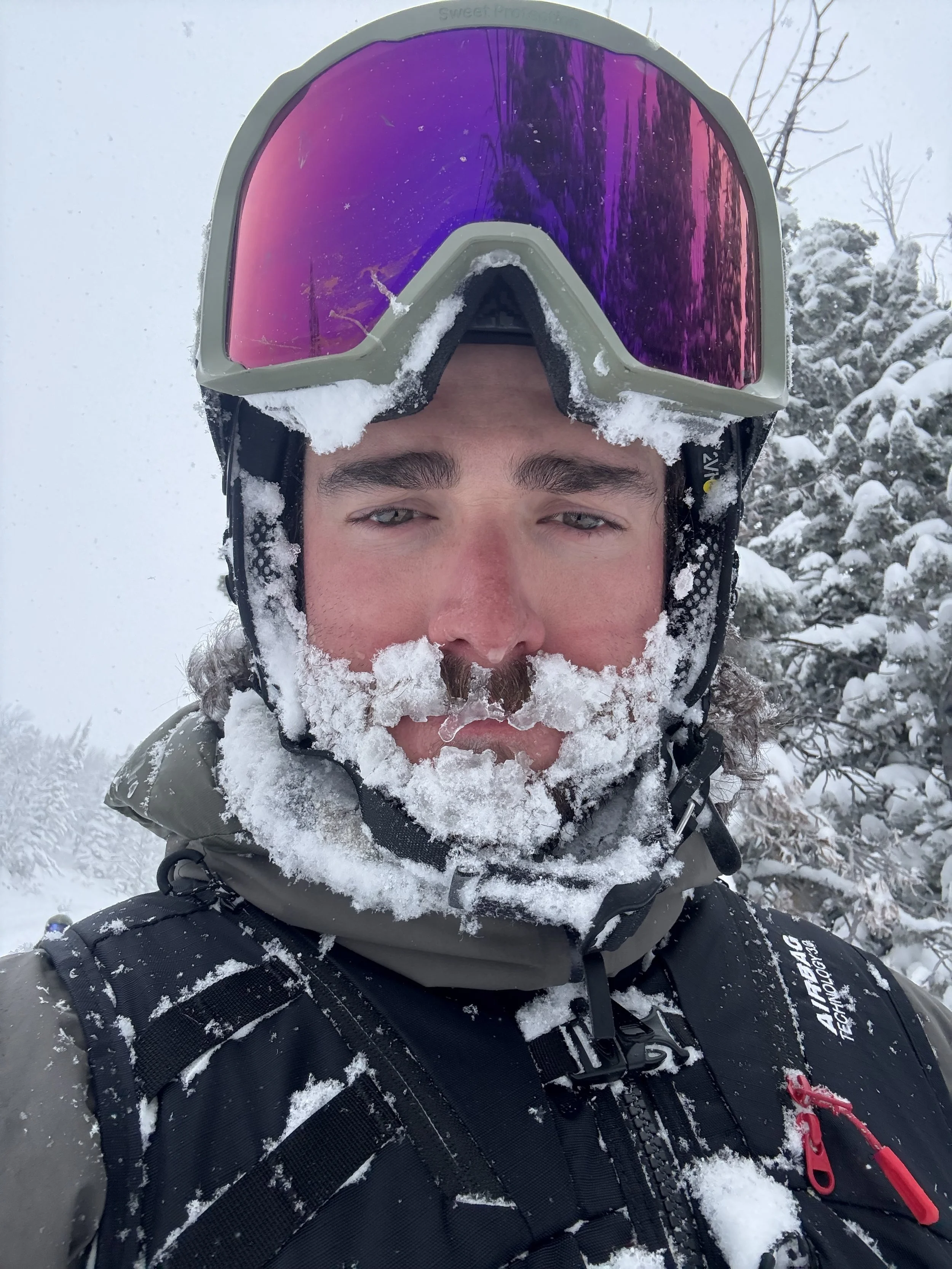 A man in snow gear with snow on his face and clothes, wearing a helmet and goggles with a snowy outdoor background.