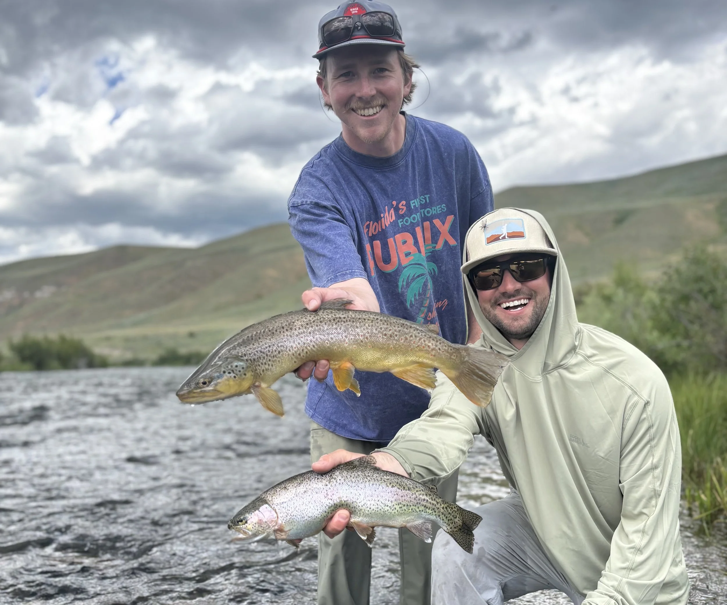 Two men fishing in a river during the day, holding up two fish they caught, with landscape hills and cloudy sky in the background.