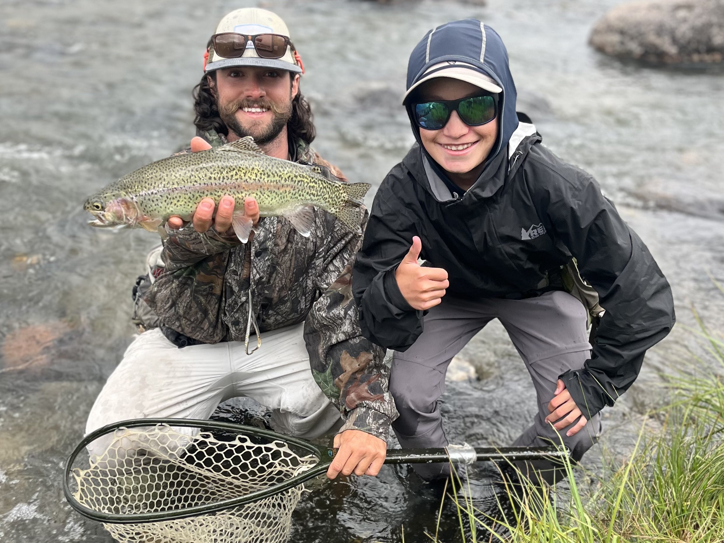 Two people, a man and a woman, are kneeling in a shallow river, holding a large rainbow trout fish. The man is wearing a camouflage jacket and a hat, while the woman is dressed in a black waterproof jacket and gray pants. Both are wearing sunglasses and are smiling, with the woman giving a thumbs-up gesture.