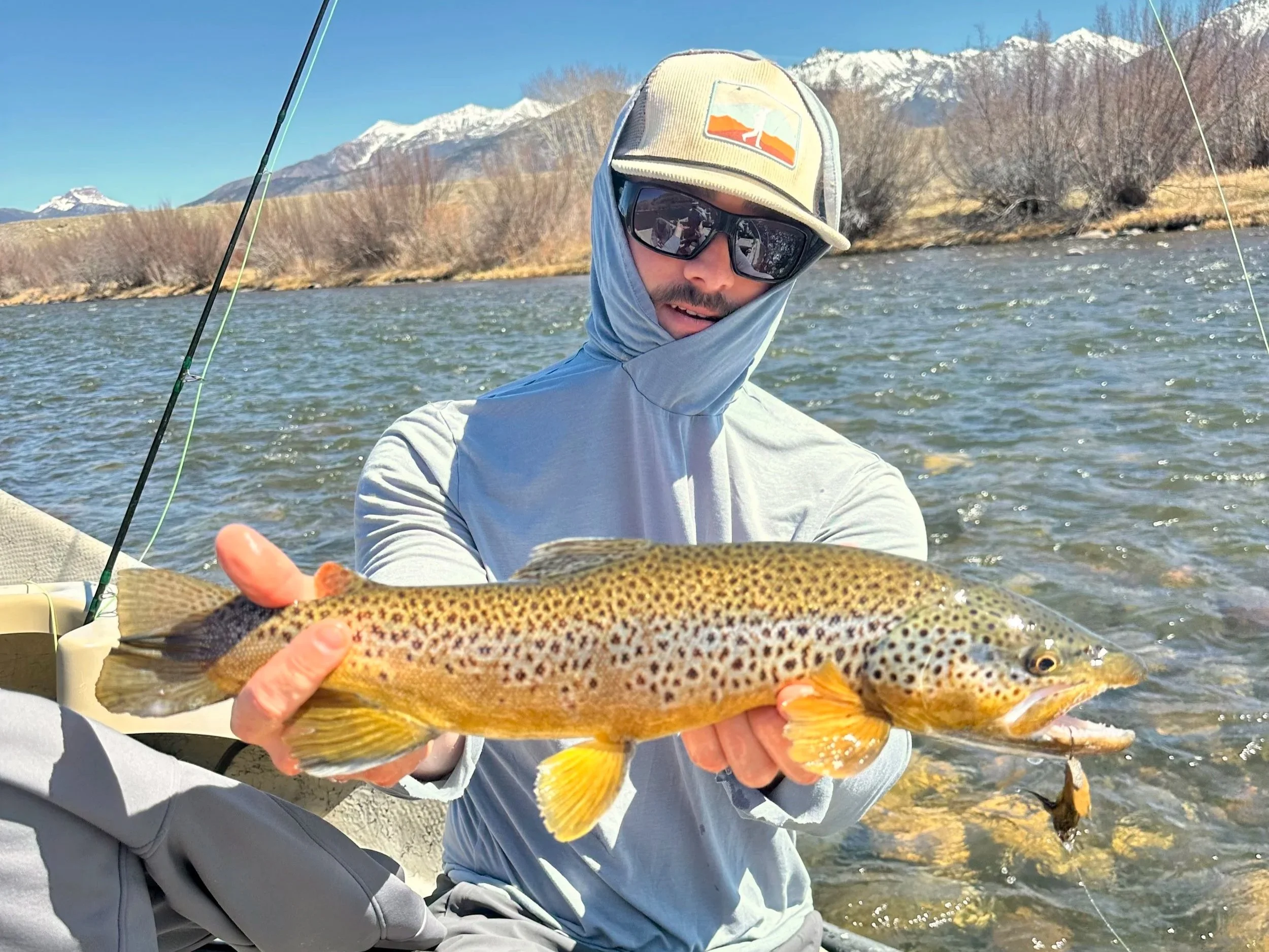 A man wearing sunglasses, a cap, and a light hoodie holding a large rainbow trout near a river with a mountainous landscape in the background.