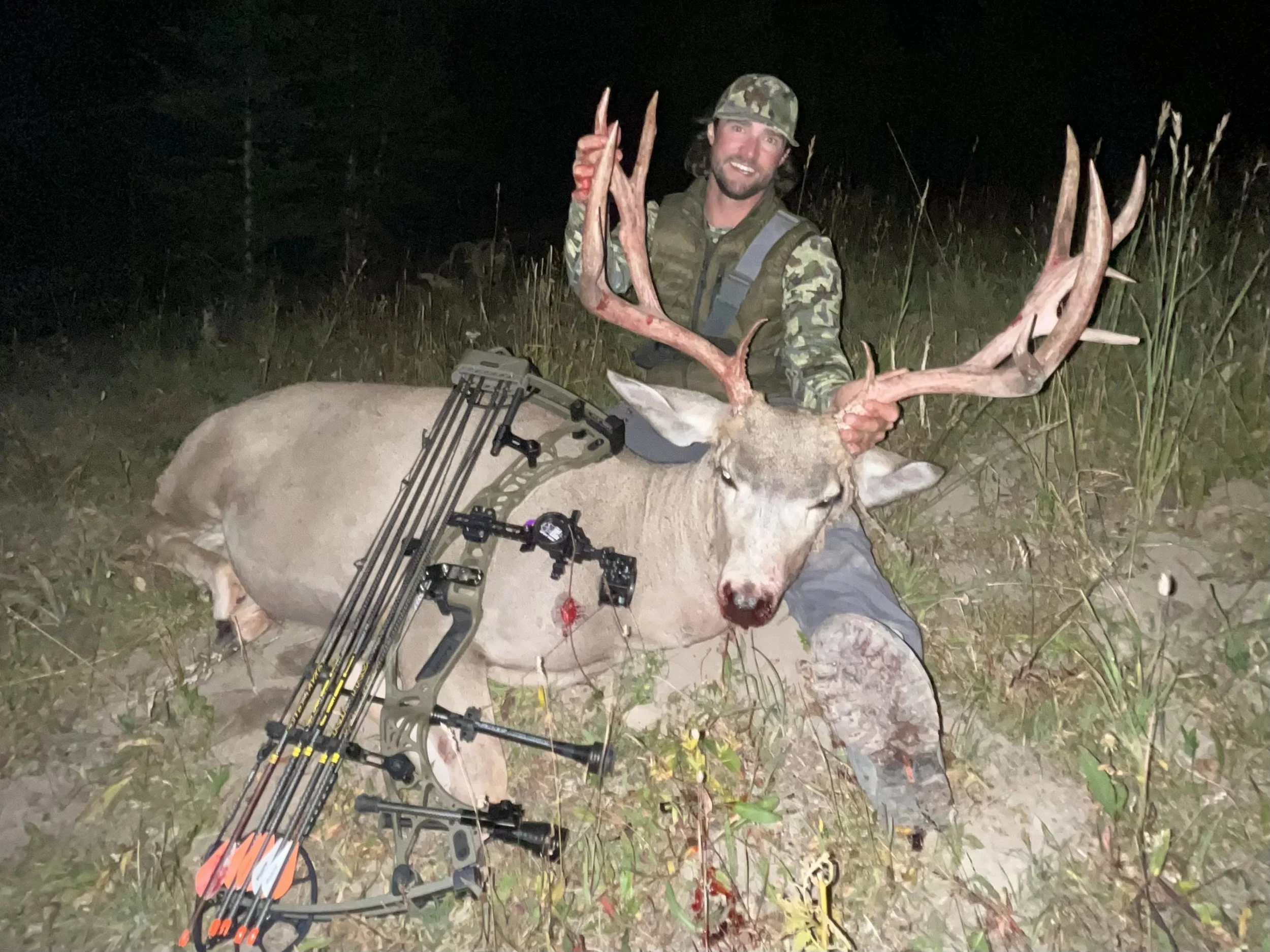 A man in camouflage clothing and a cap holding antlers of a large harvested deer with a bow and arrows lying on the deer in a nighttime outdoor setting.