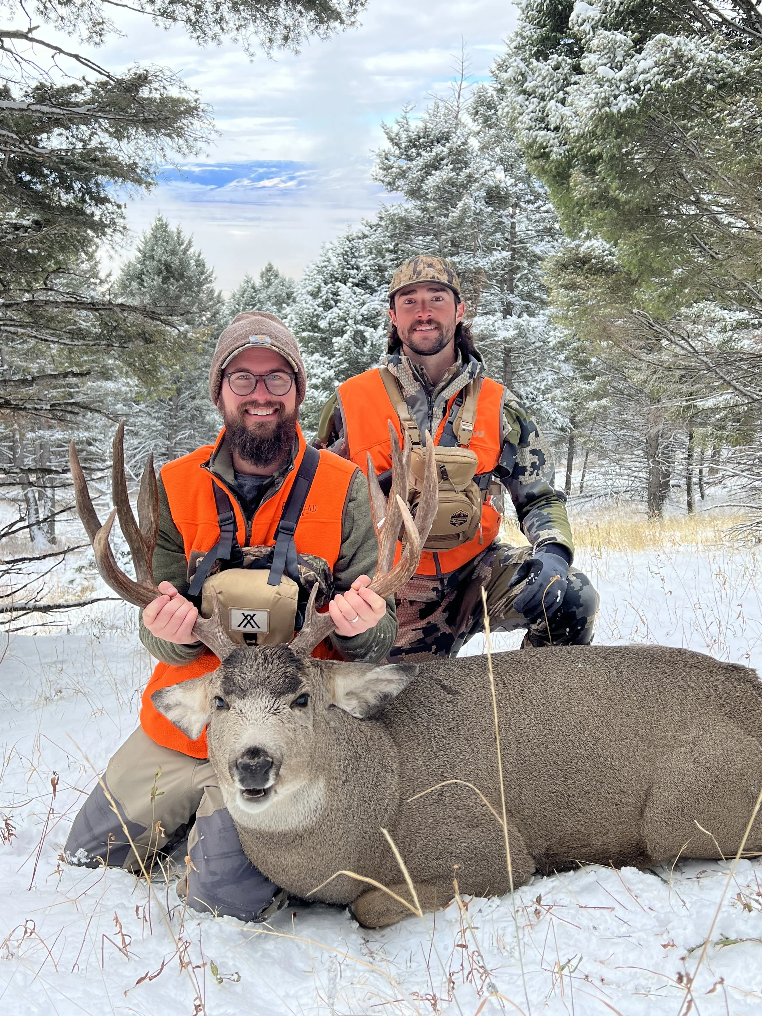 Two hunters kneeling in snow-covered forest with a large deer they have hunted, both wearing orange hunting vests, holding the deer's antlers, smiling.