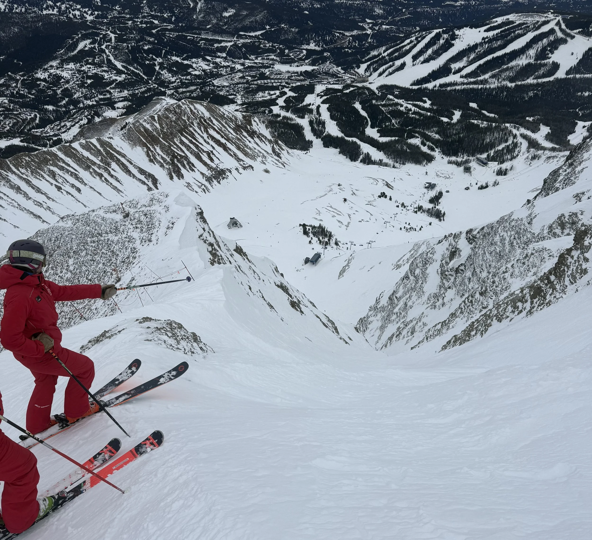 A skier dressed in red gear skiing down a snowy mountain slope with a view of a valley and distant mountains in the background.