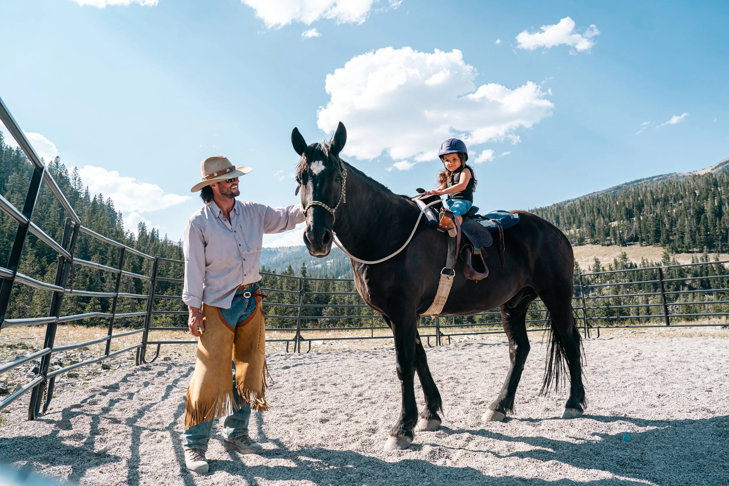 A man helping a young girl ride a black horse in an outdoor arena with mountains and trees in the background under a partly cloudy sky.