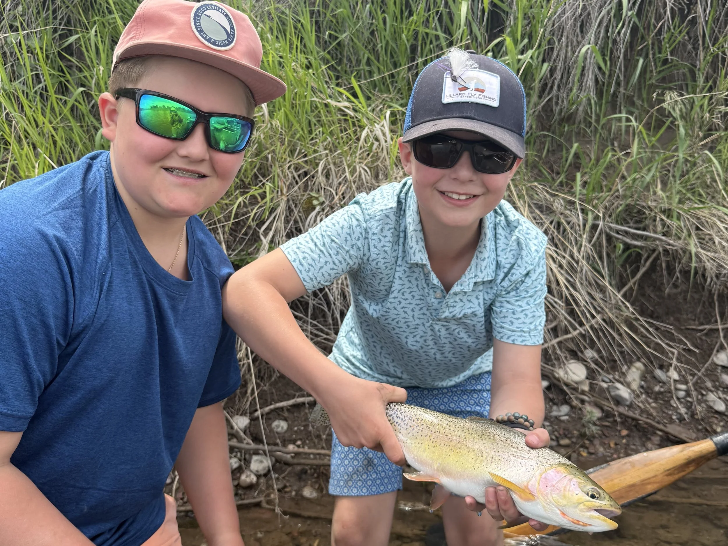 Two boys, wearing sunglasses and hats, are outdoors near a creek, holding a large fish they caught.