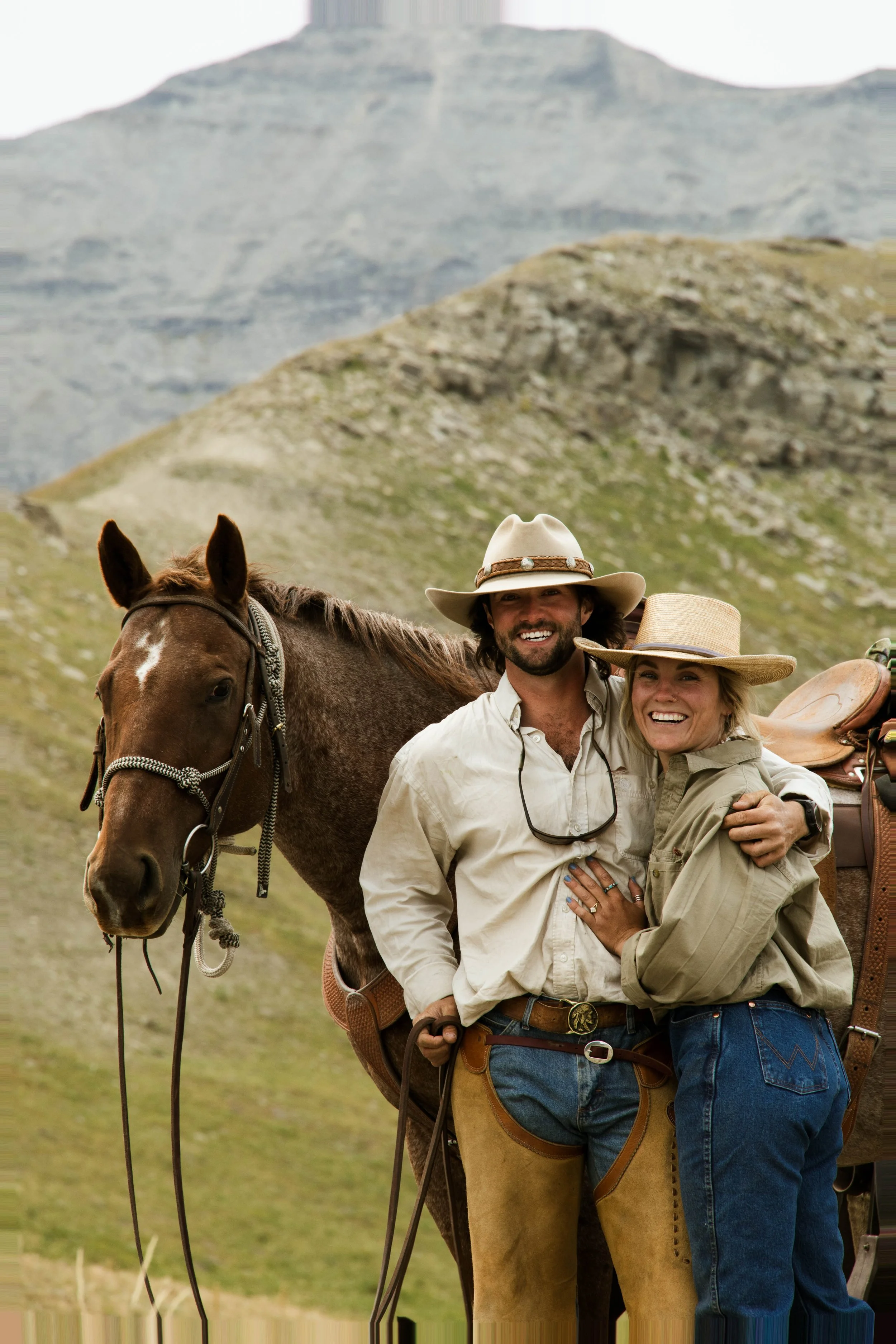 A man and woman embracing and smiling outdoors with a horse and mountainous landscape in the background.
