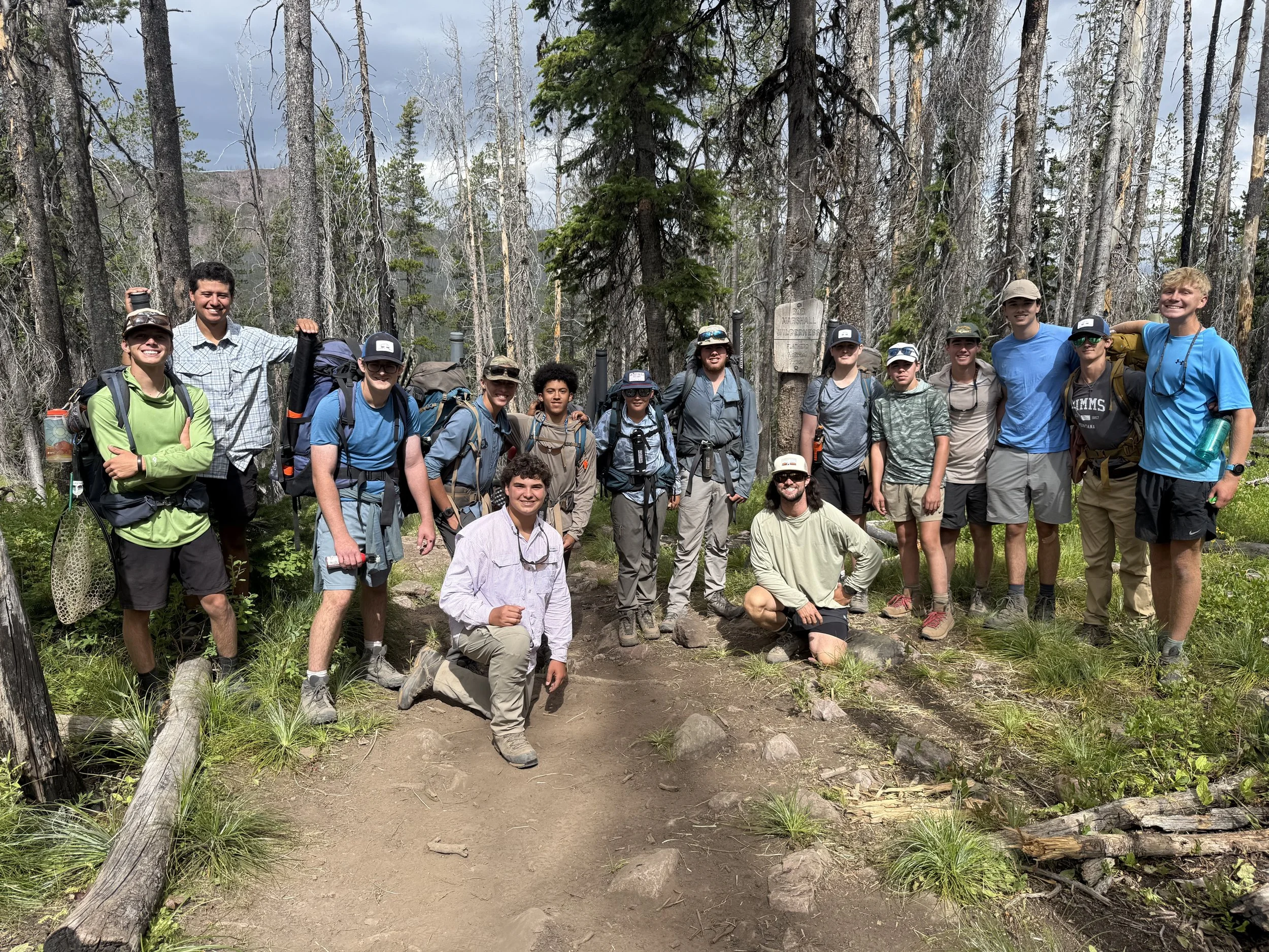Group of 16 young hikers, wearing backpacks and outdoor gear, posing on a forest trail with trees and greenery in the background.