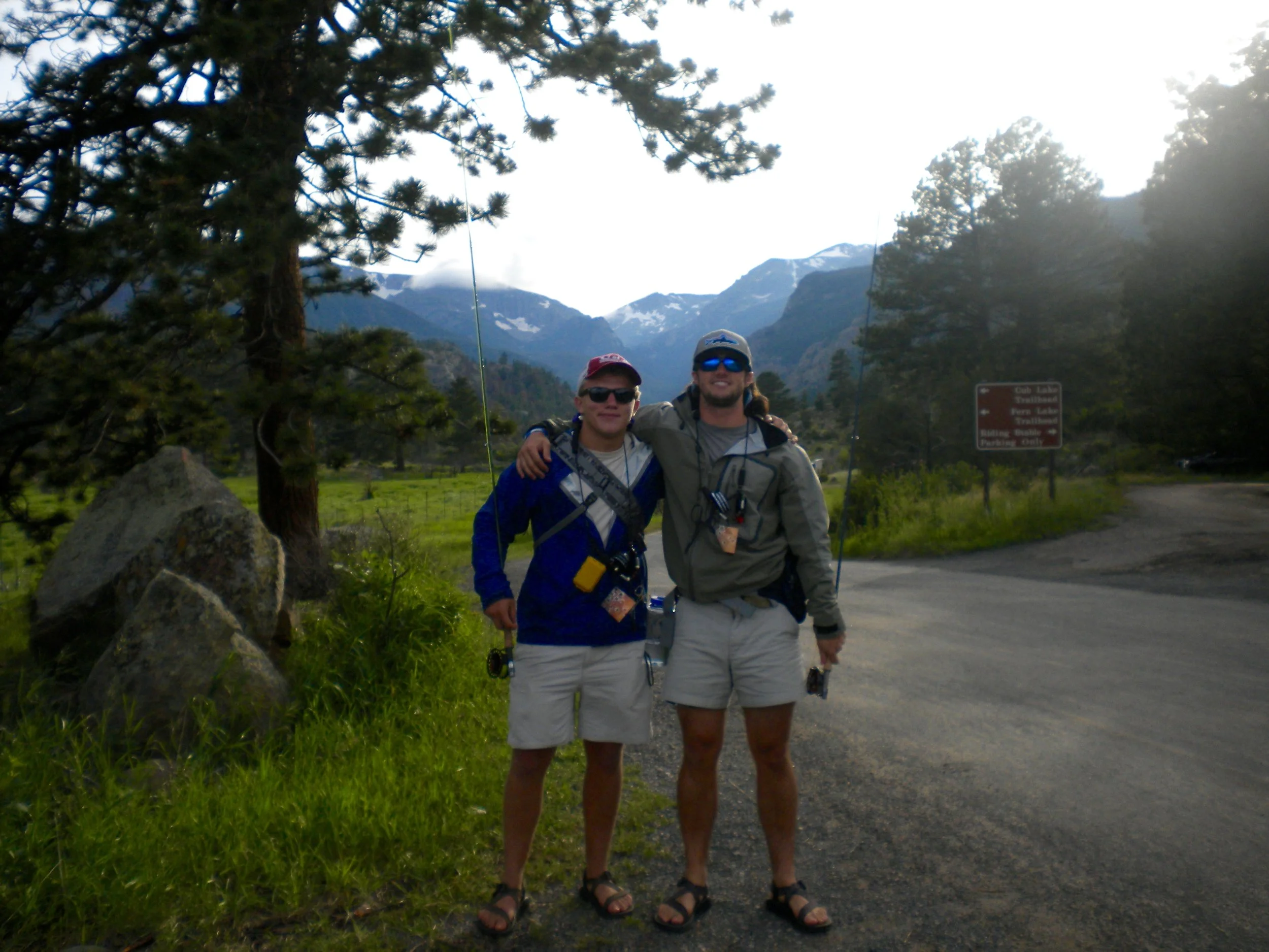 Two men in hiking gear standing outdoors on a mountain trail with trees, rocks, and mountains in the background.