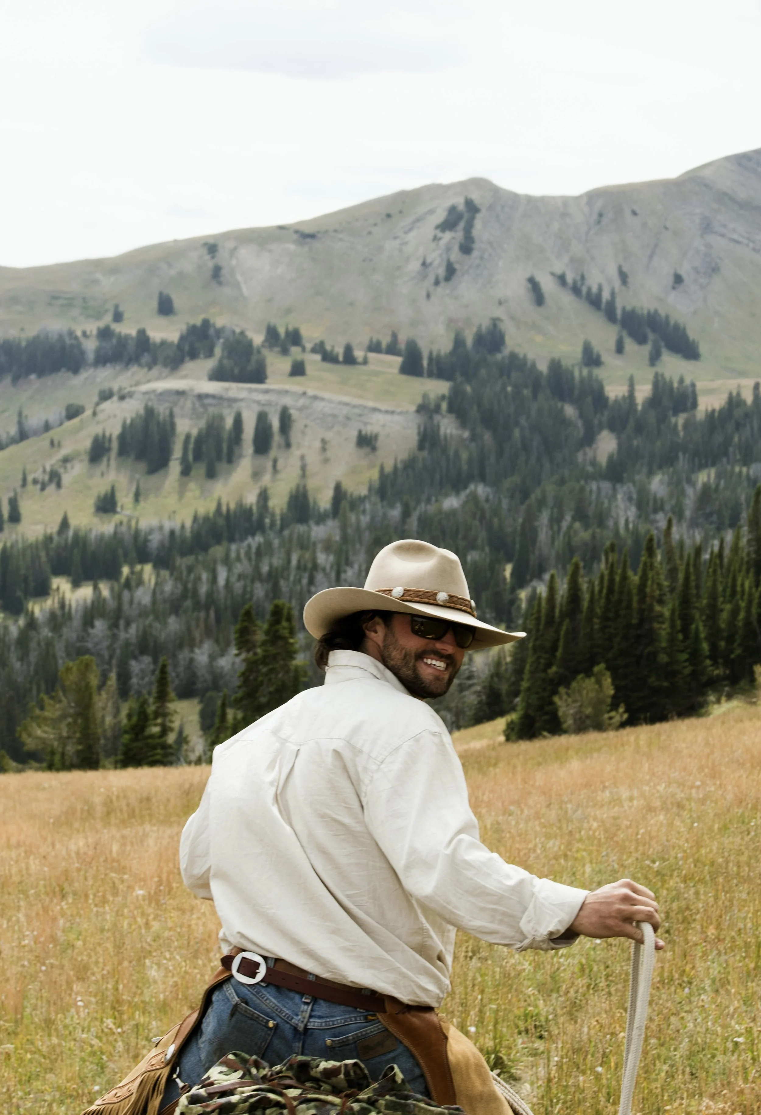 A man wearing a cowboy hat, sunglasses, and a beige shirt, smiling and walking through a grassy field with a mountainous forest landscape in the background.