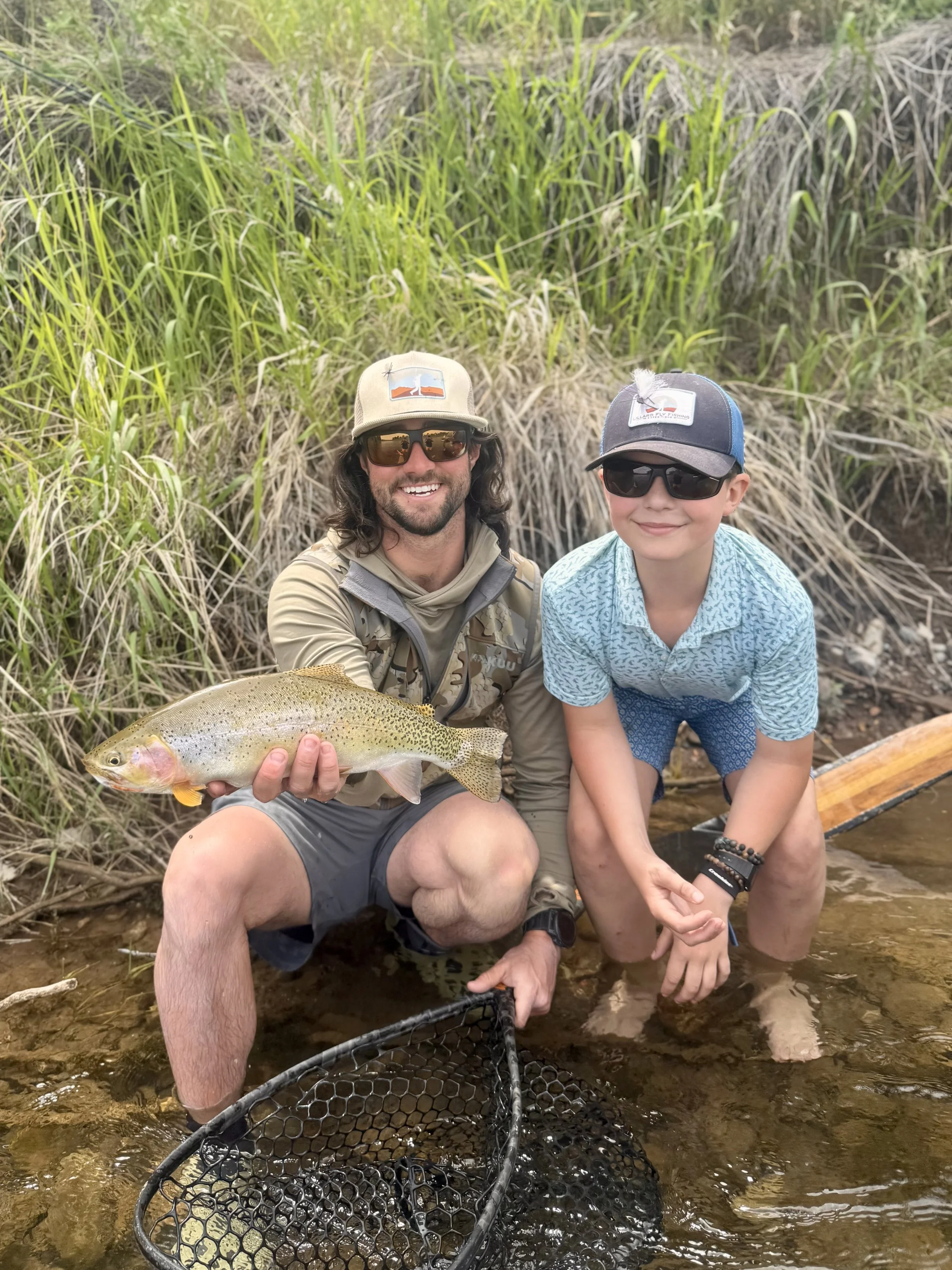 A man and a boy kneeling in a stream, holding a large rainbow trout, with tall grass and reeds in the background.