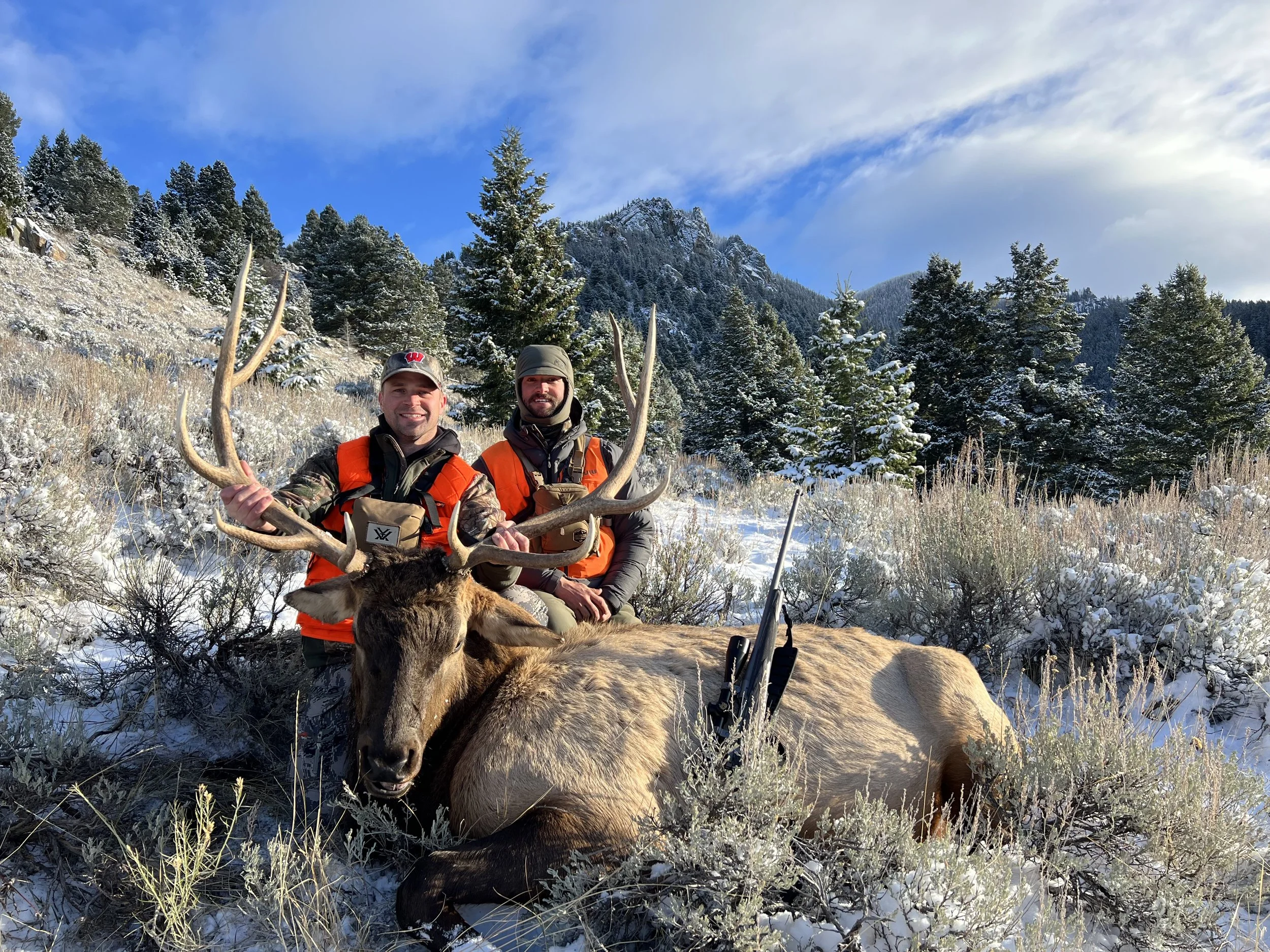 Two hunters in orange vests pose with a large buck with antlers in a snowy forested landscape.