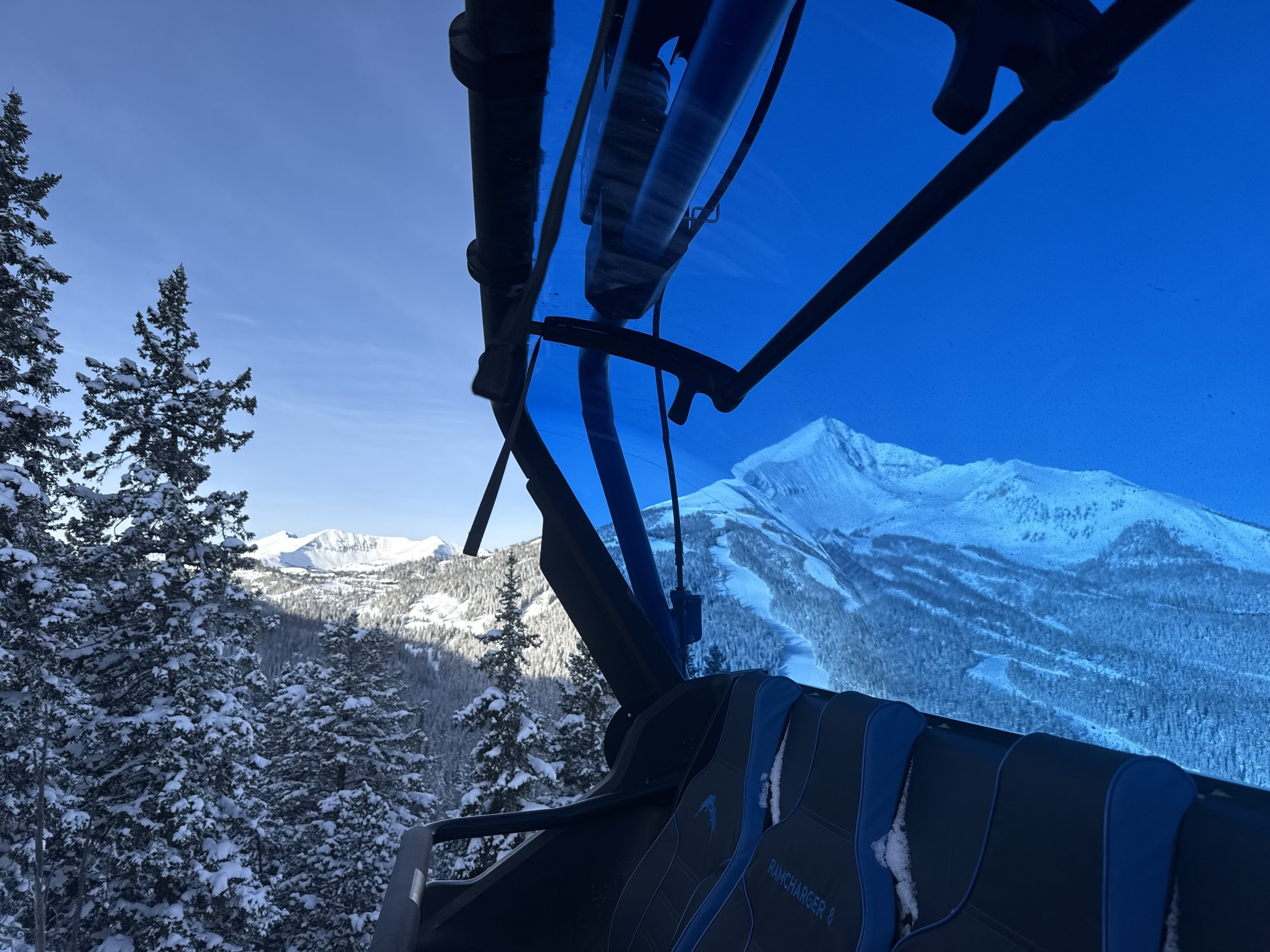 Inside a snowcat vehicle with a view of snow-covered pine trees and mountain peaks through a window.