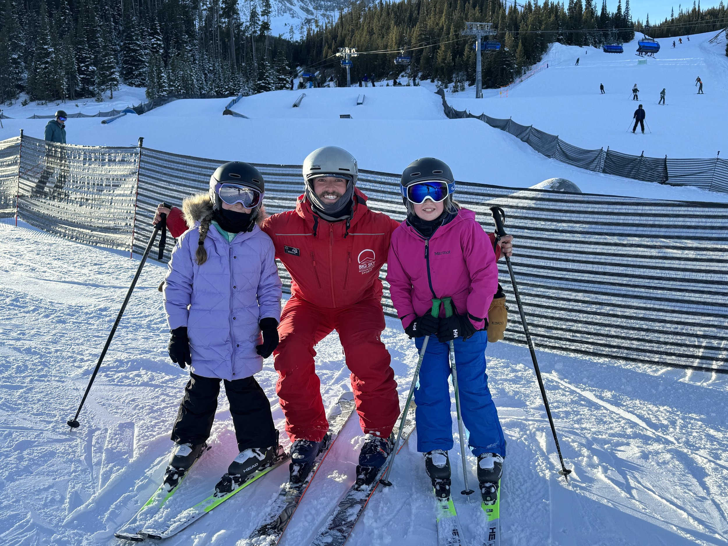 Three people in ski gear posing on a snowy mountain slope, with skiers and ski lifts in the background.