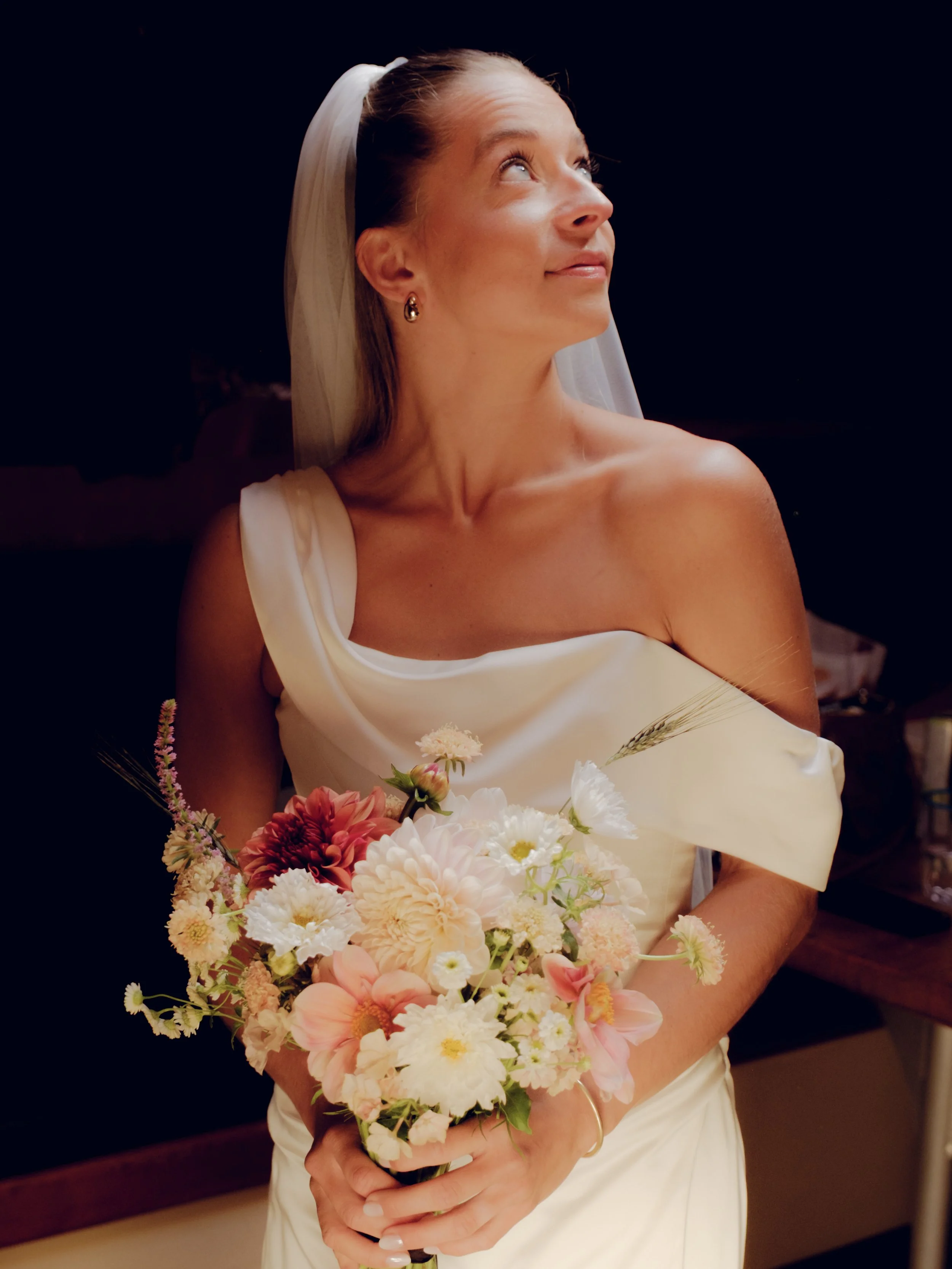 A bride in a white wedding dress holding a bouquet of pink, white, and peach flowers, looking upward with a gentle smile.