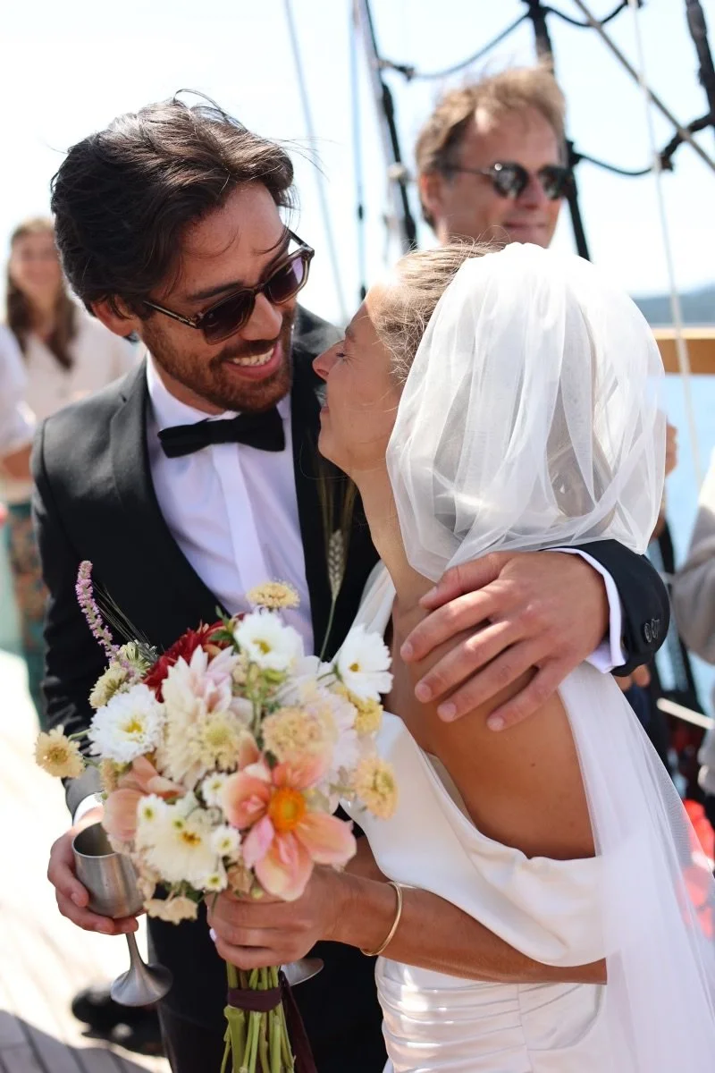 A couple celebrating their wedding day outdoors, with the groom wearing sunglasses and a tuxedo, and the bride wearing a veil and holding a bouquet of flowers, embracing and smiling.