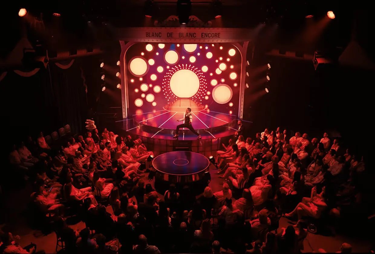 A performer on stage at a circus or show, surrounded by an audience, with a large, colorful backdrop featuring glowing circles and a circular platform in the center.