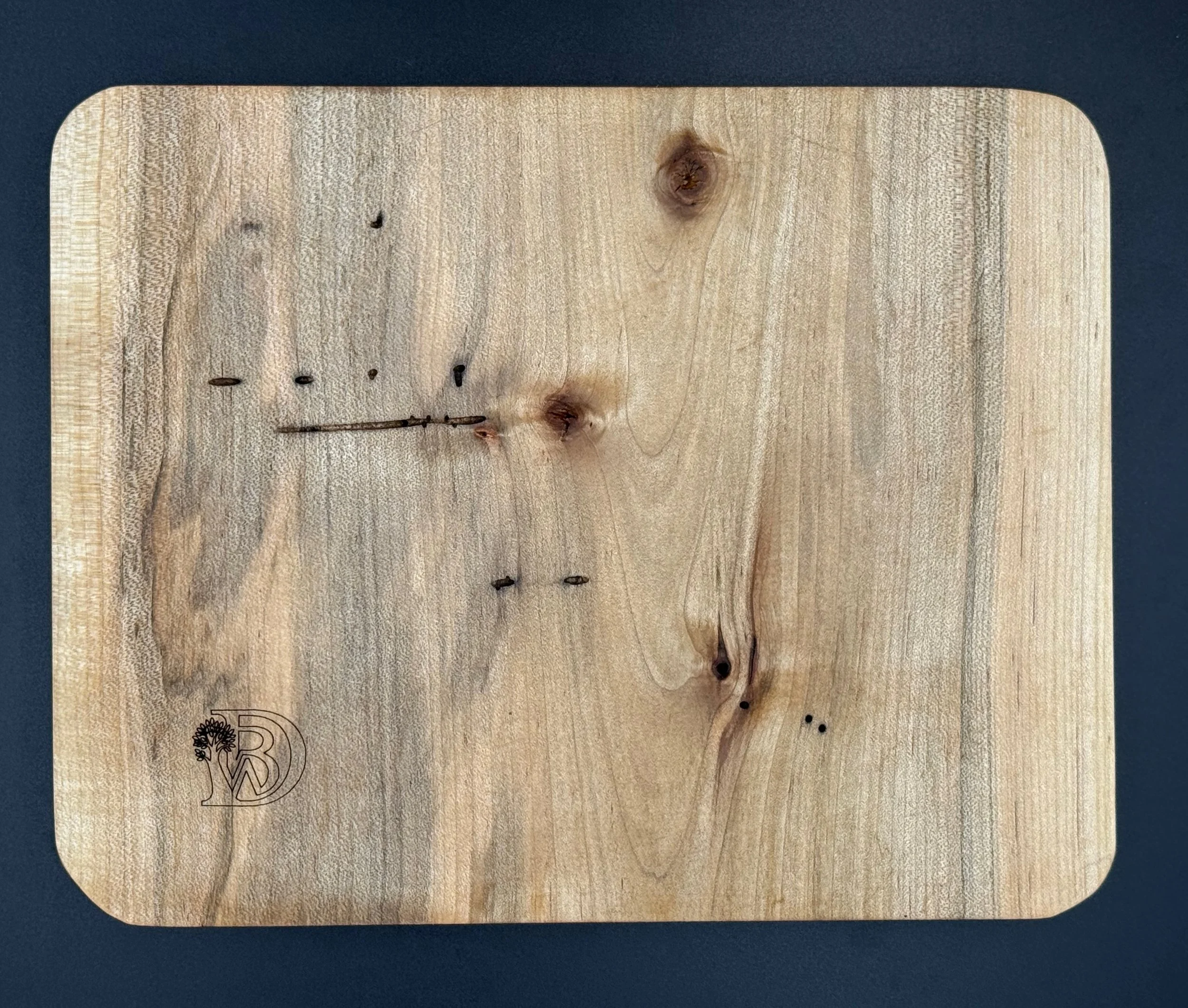 A wooden panel with the initials 'BD' and a small tree logo engraved in the lower left corner, placed on a dark surface.