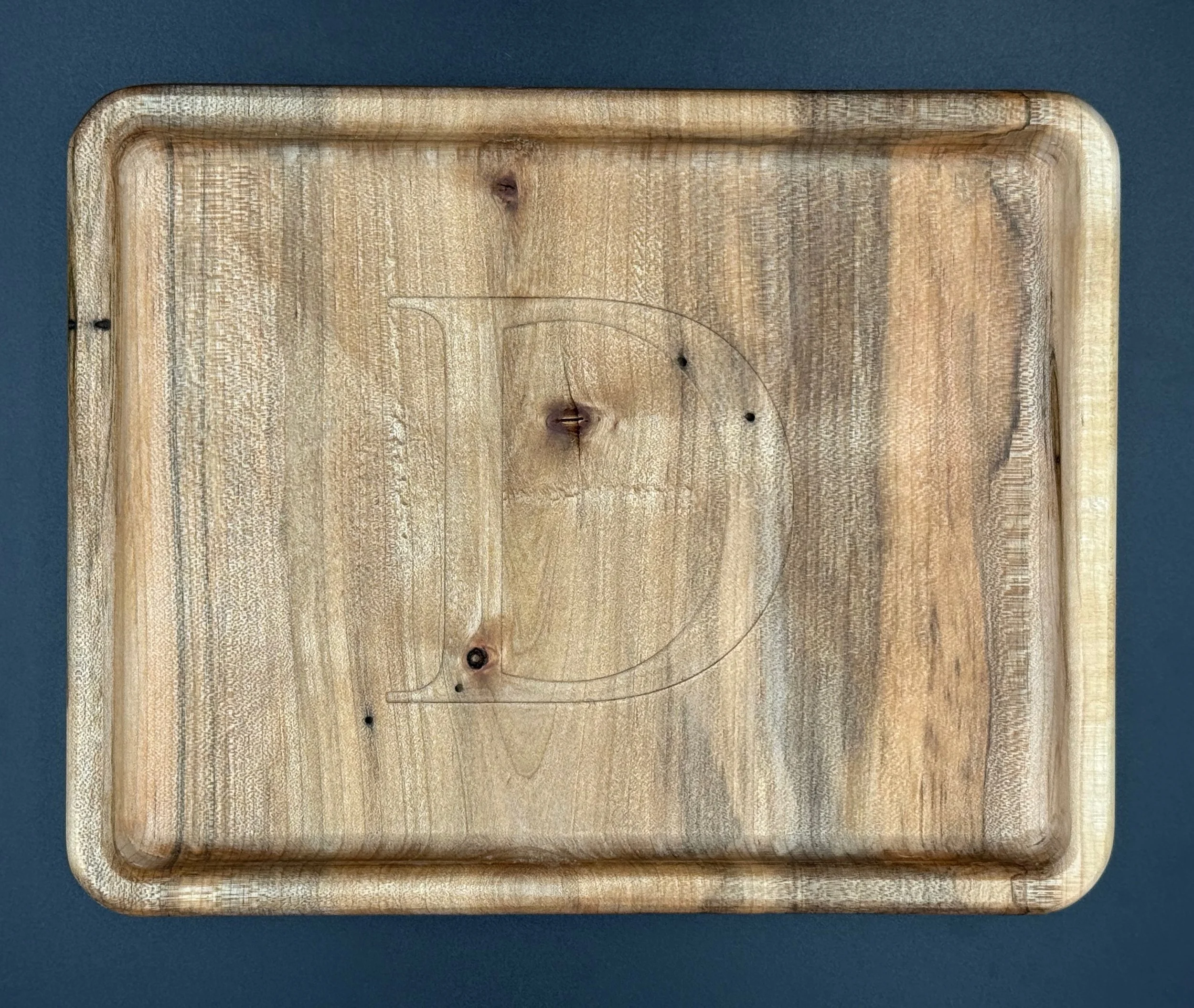 Wooden cutting board with carved outline in the shape of the letter D, placed on a dark surface.