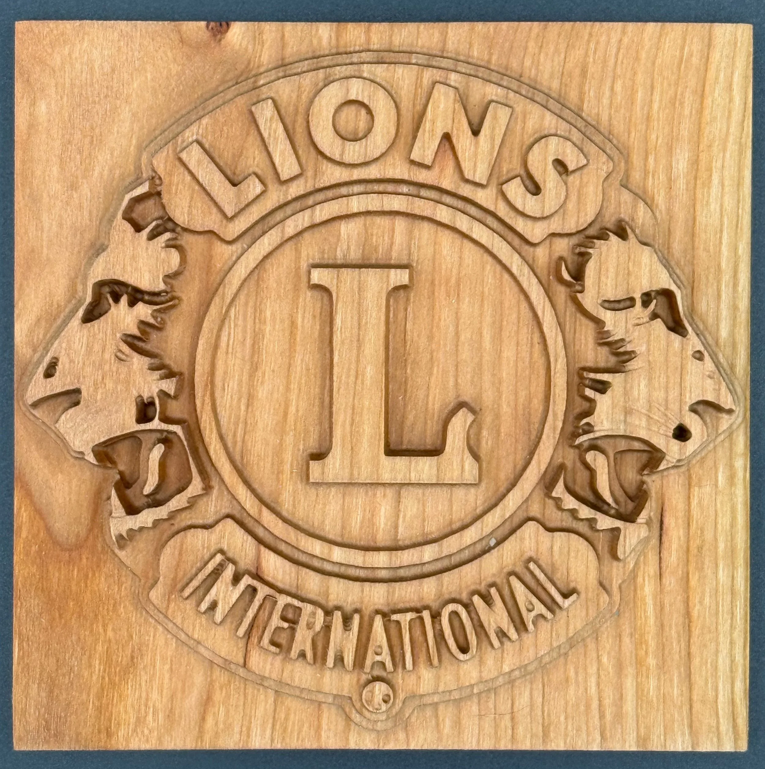 Wooden plaque engraved with the words "LIONS INTERNATIONAL" and a circular logo with a large letter 'L' in the center, surrounded by two lion silhouettes facing away from each other.