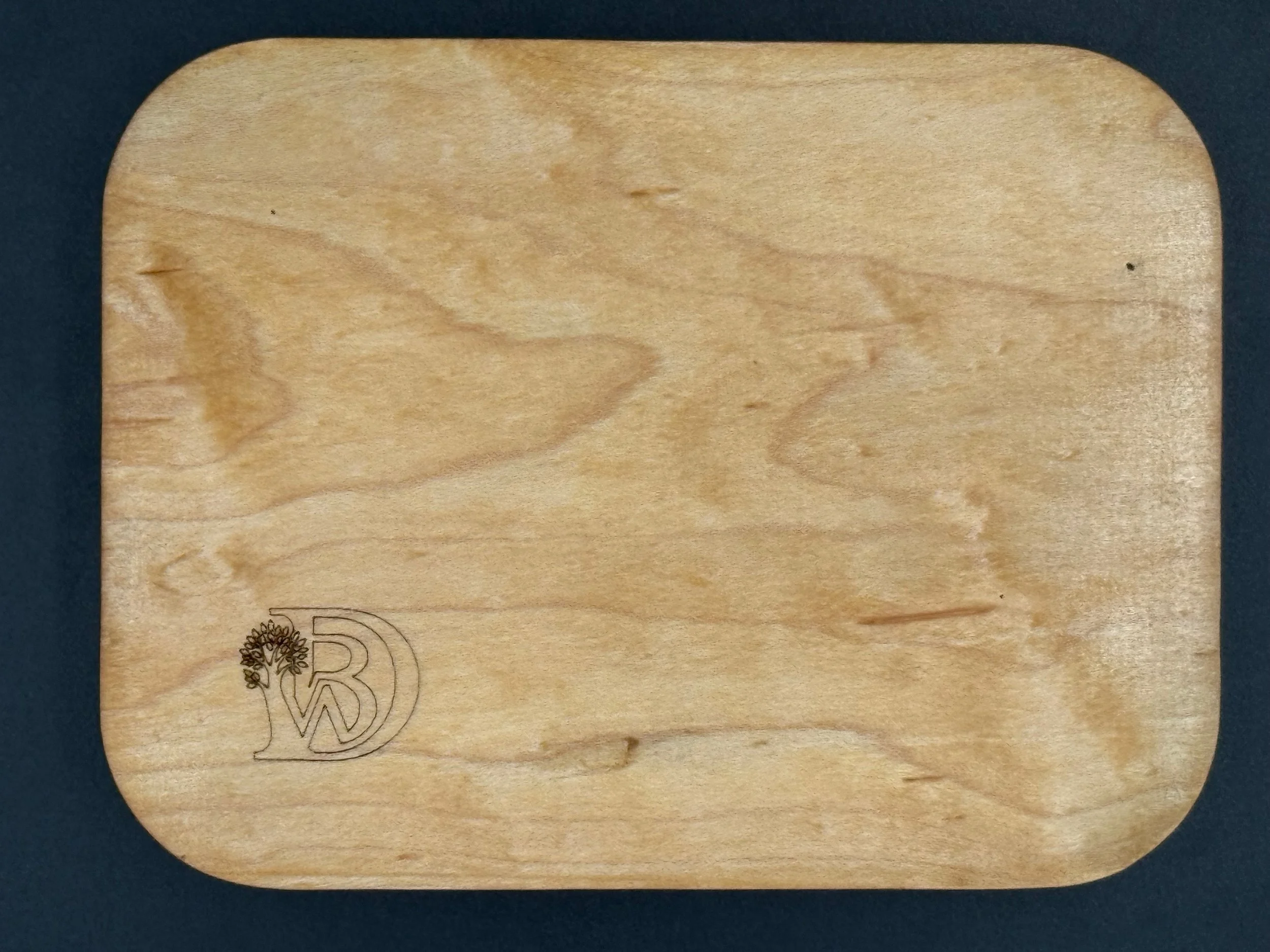 A square wooden cutting board with rounded corners, featuring a laser-engraved logo of a stylized tree leaves and the letters 'W' and 'D'.