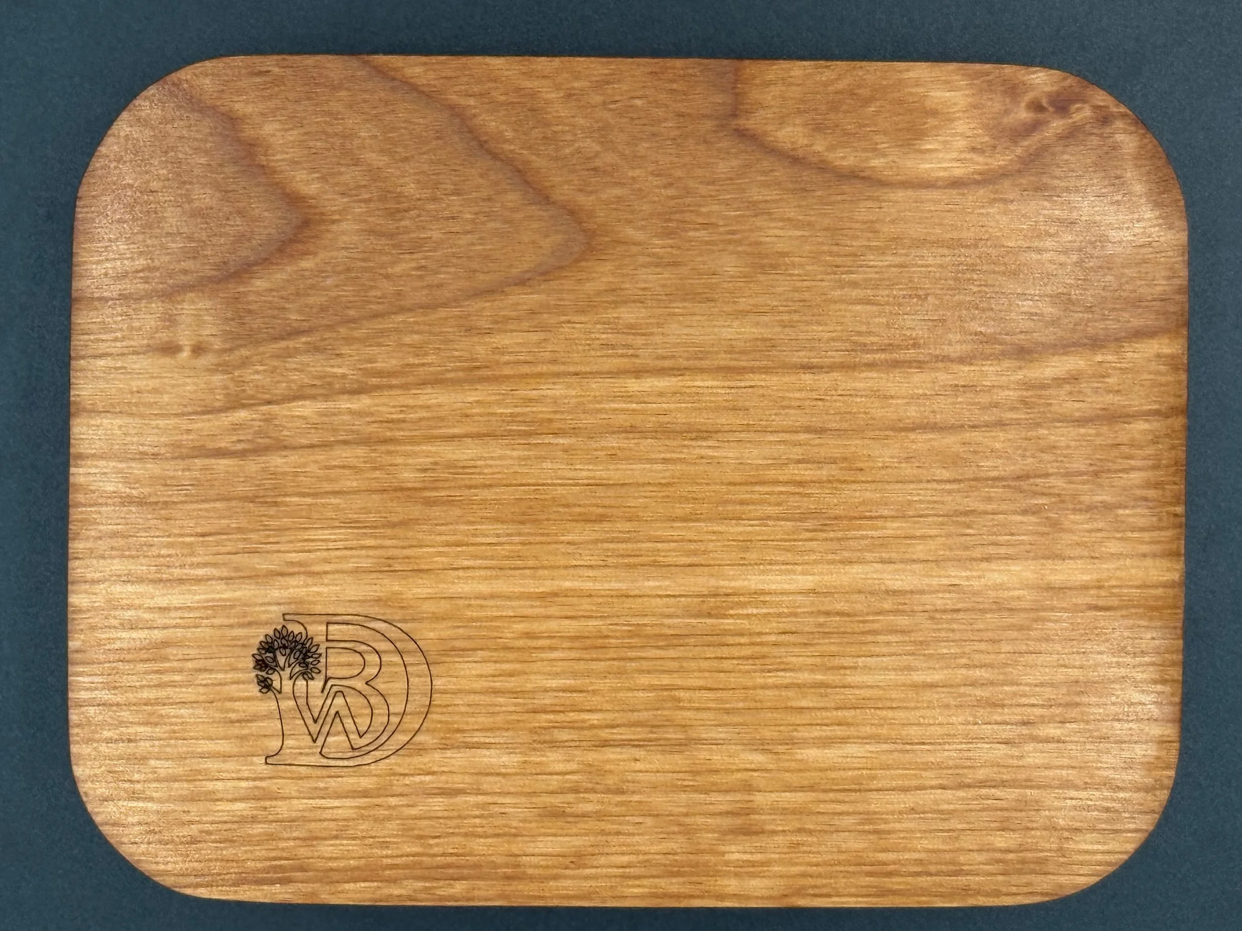A rectangular wooden cutting board with rounded edges and a small engraved logo of a tree and the letter 'W' in the bottom left corner.