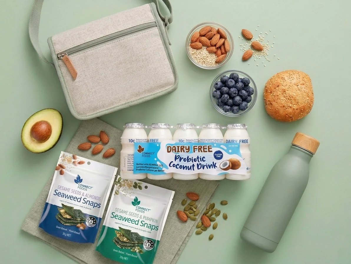 A flat lay of a light gray shoulder bag, halved avocado, pack of seaweed snacks, probiotic coconut drink, blueberries, almonds, pumpkin seeds, a sesame seed bun, a water bottle, and small bowls of almonds and blueberries on a light green background.
