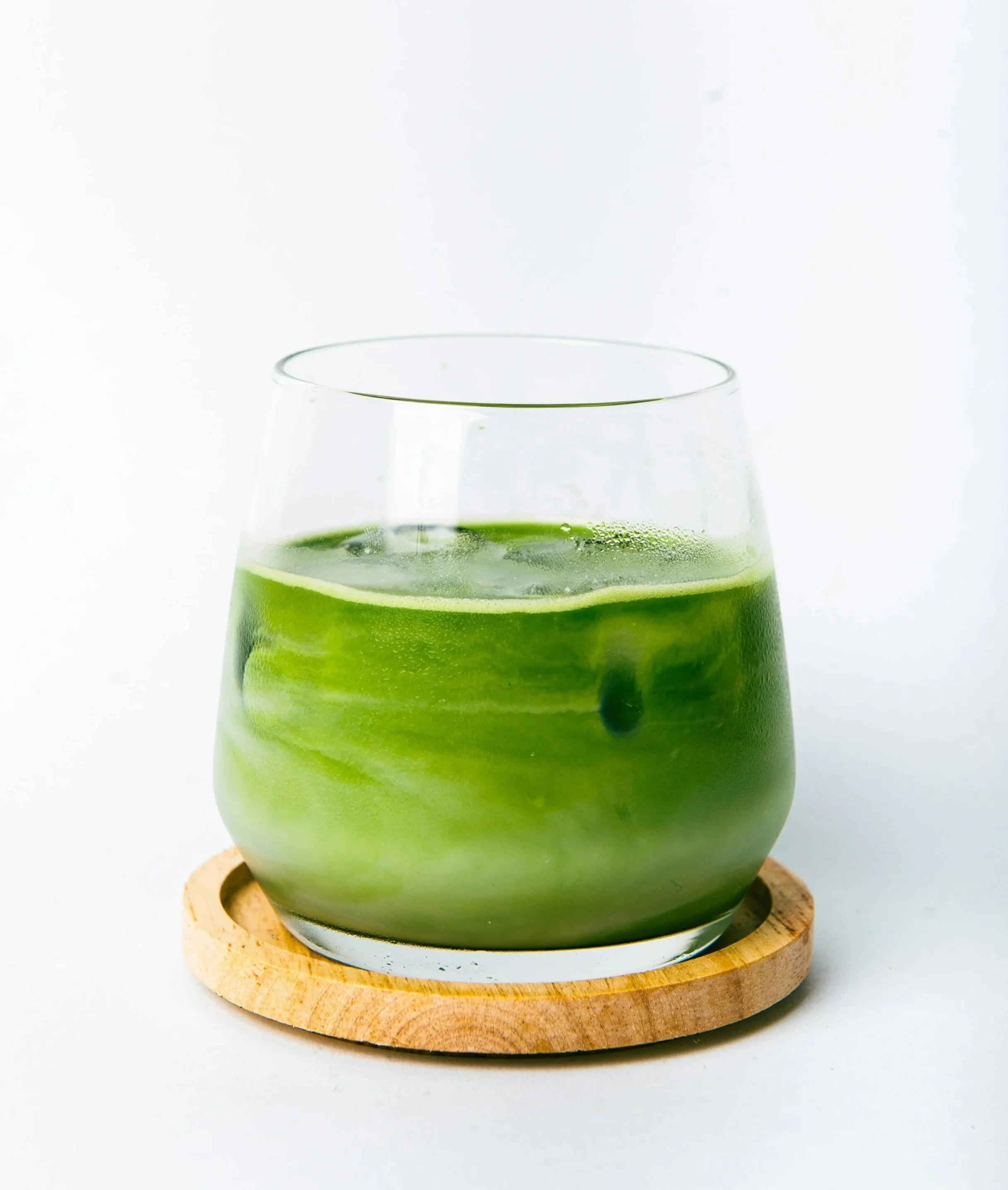 A glass of green juice with a slice of cucumber garnishing it, placed on a round wooden coaster against a plain white background.