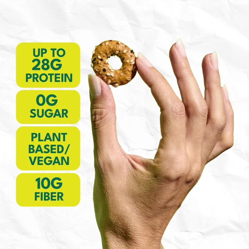 A hand holding a small, round, multigrain snack with text highlighting its nutrition facts: up to 28 grams of protein, 0 grams of sugar, plant-based/vegan, and 10 grams of fiber.