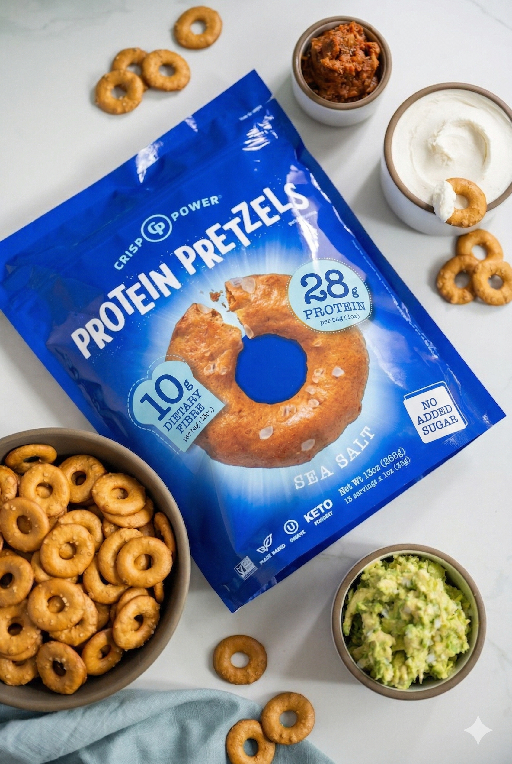 Bag of CRISP POWER Protein Pretzels in Sea Salt flavor with bowls of pretzels, guacamole, salsa, and yogurt.