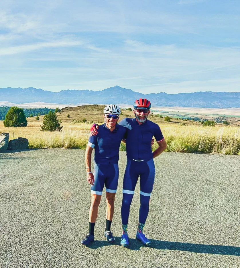 Cycling coach Woody standing with another cyclist with arms around each other in front of pretty view of brush and mountains.