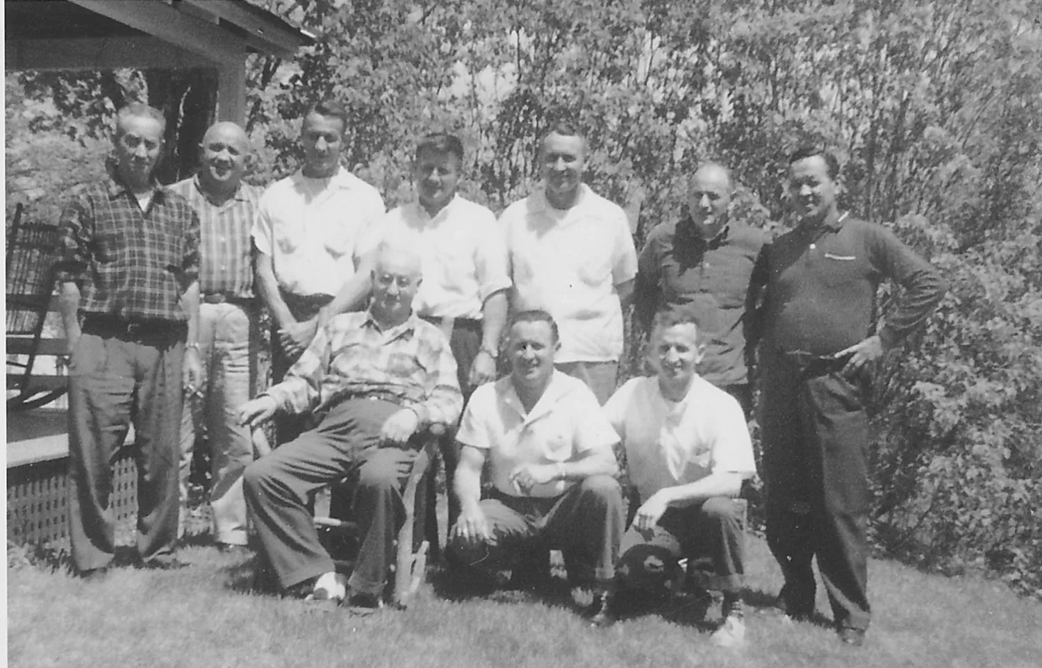 A black-and-white photo of eleven men outdoors, some standing and some sitting, in front of trees and a house porch. The men are dressed in casual shirts and pants, and are smiling at the camera.