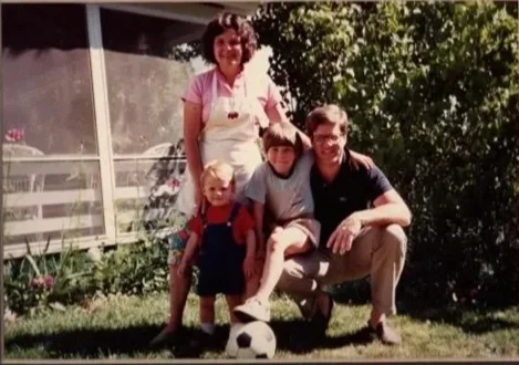 Family photo of four people outdoors with a dog and a soccer ball, standing on grass in front of a house with plants and trees.