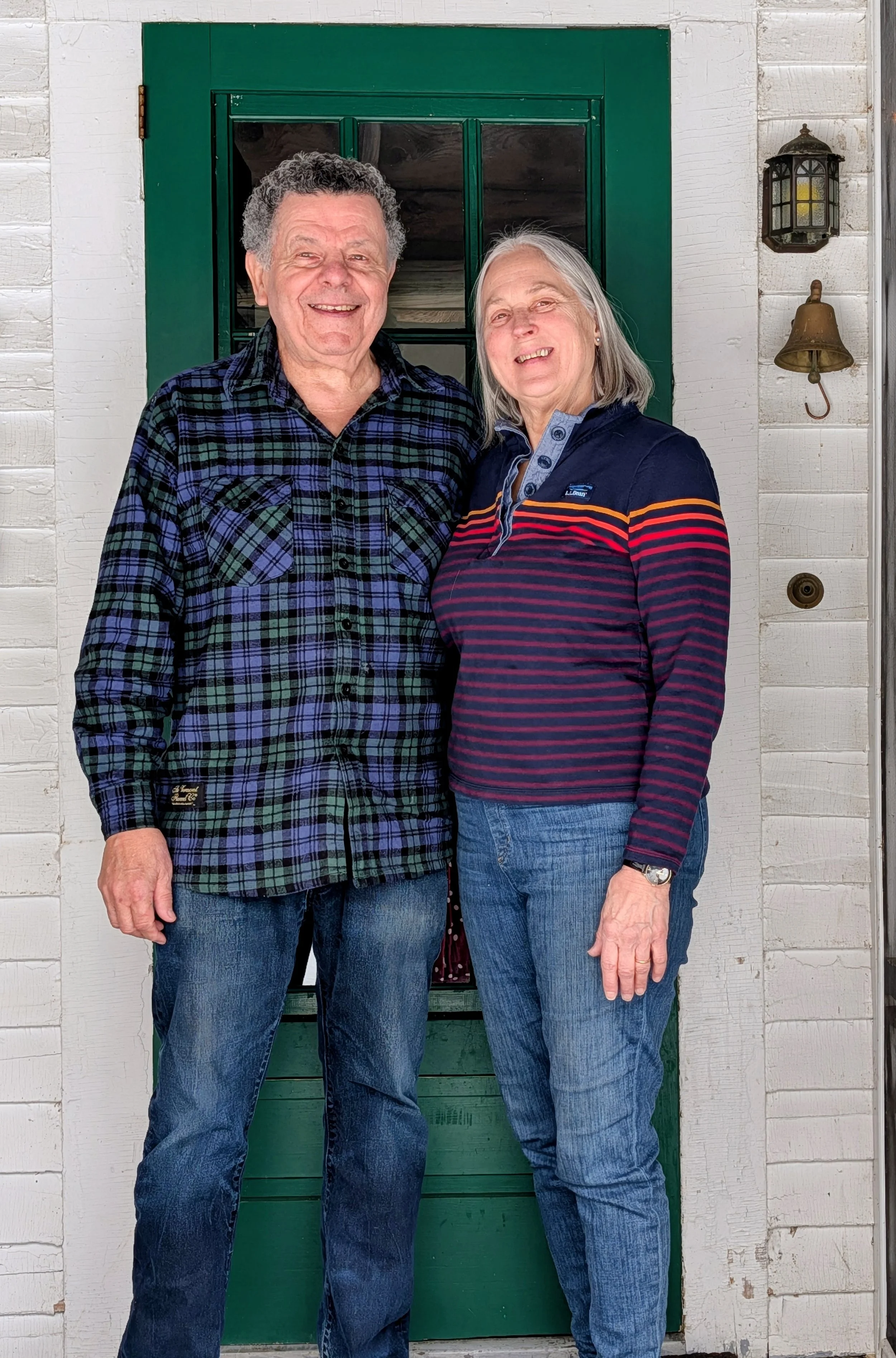 A smiling elderly man and woman standing together in front of a green door, outside a white wooden house, with a lantern and a bell on the wall.