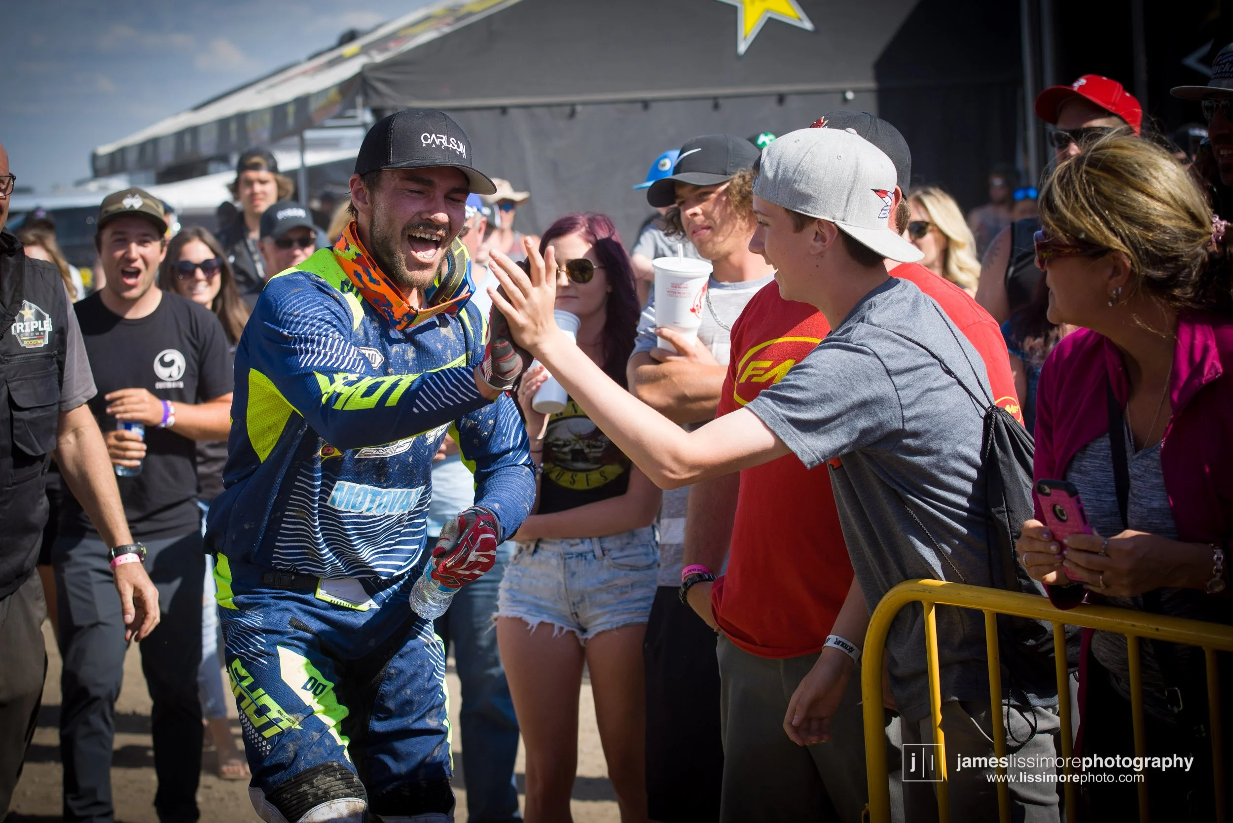 A motocross rider in a blue and yellow racing suit is giving a high five to a fan in a grey t-shirt and white cap, surrounded by other fans behind a yellow barrier, with a black tent and cloudy sky in the background.