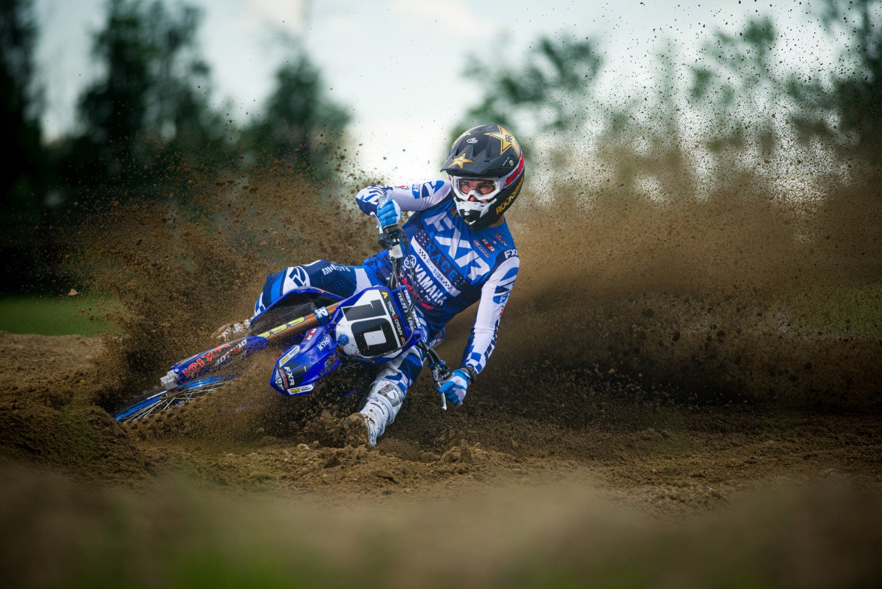 Motocross rider in blue gear and helmet riding a dirt bike on a dirt track, kicking up dirt and mud.